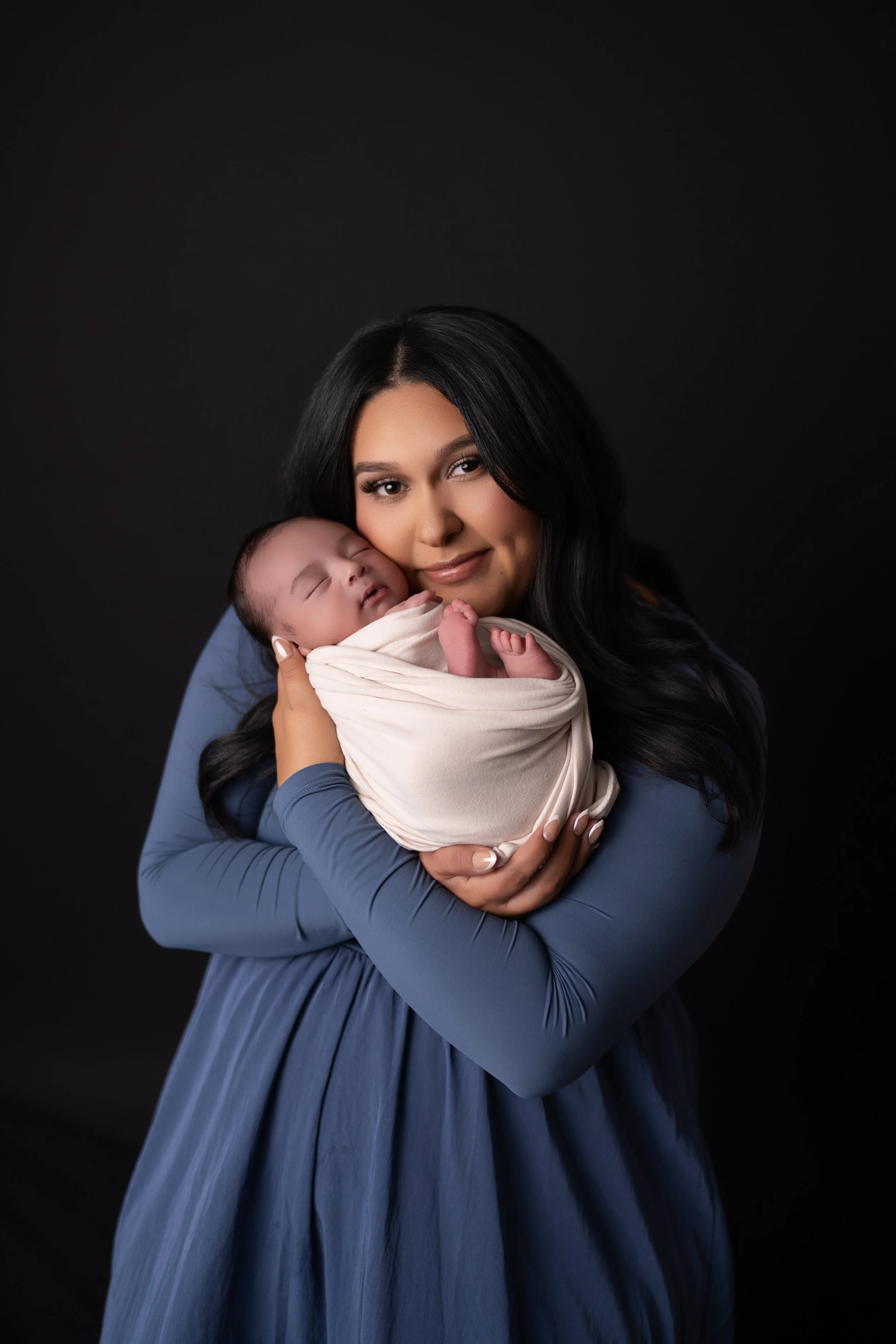 A woman with long black hair holding a sleeping baby wrapped in a beige blanket against a black background.
