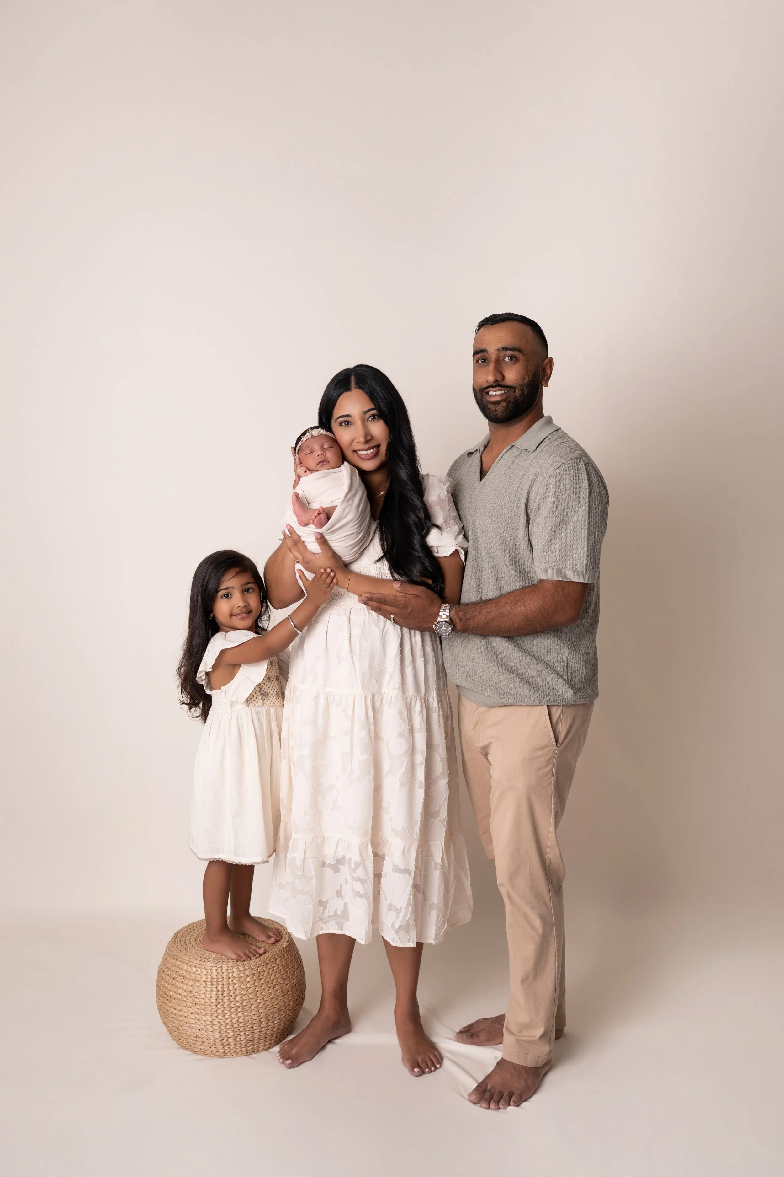 A family portrait featuring a woman, a man, an older girl, and a newborn baby against a blank white background. The woman is holding the baby, and the older girl is standing on a round woven stool, smiling, while the father stands beside them.