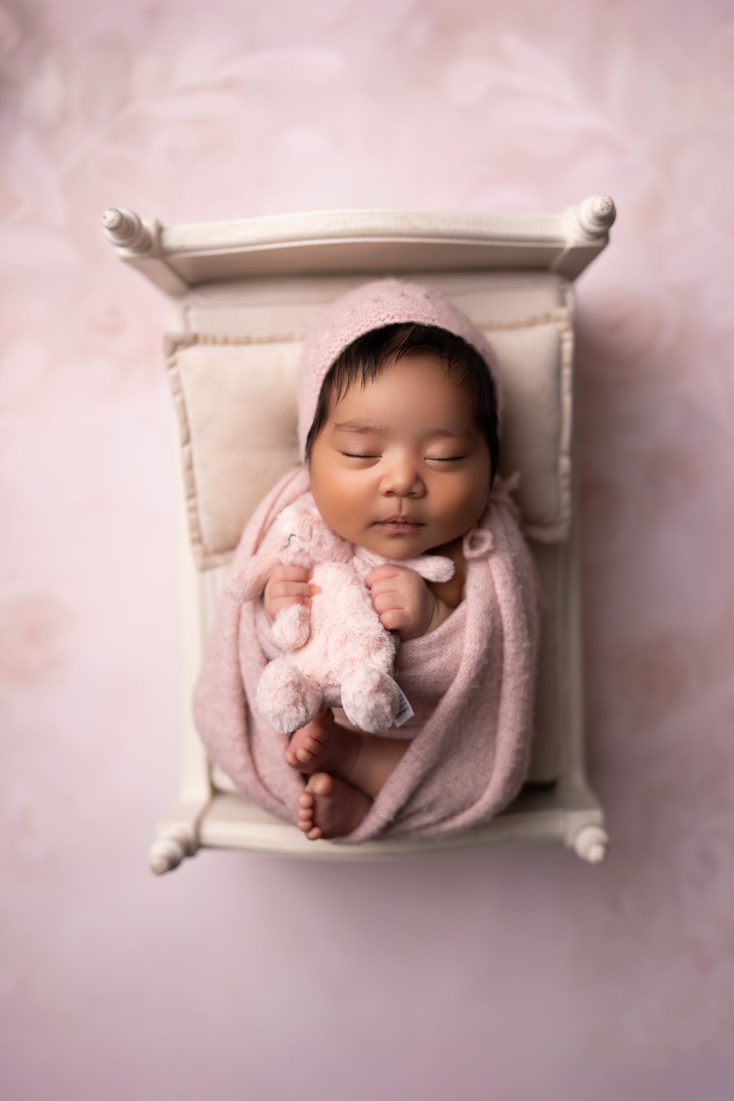 A sleeping baby wrapped in pink blanket, holding a stuffed toy, lying on a small white bed against a pink background.