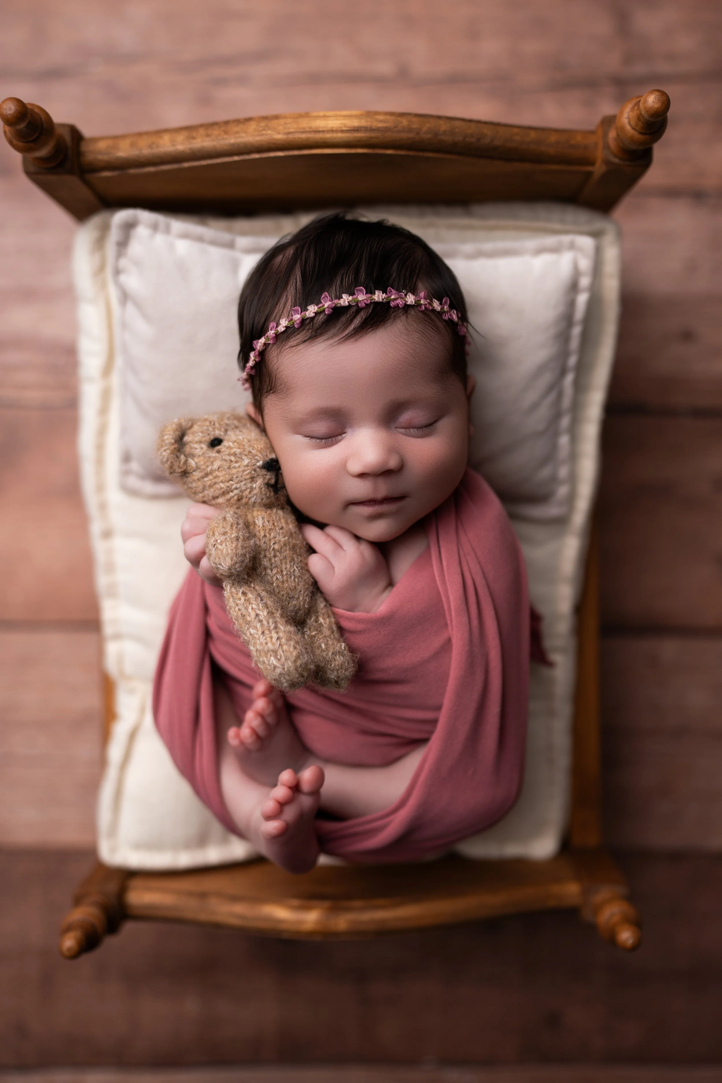 A sleeping newborn girl with dark hair, wearing a pink headband with small flowers, is swaddled in a pink cloth. She is holding a small, knitted teddy bear close to her face, lying on a small wooden bed with a beige pillow.