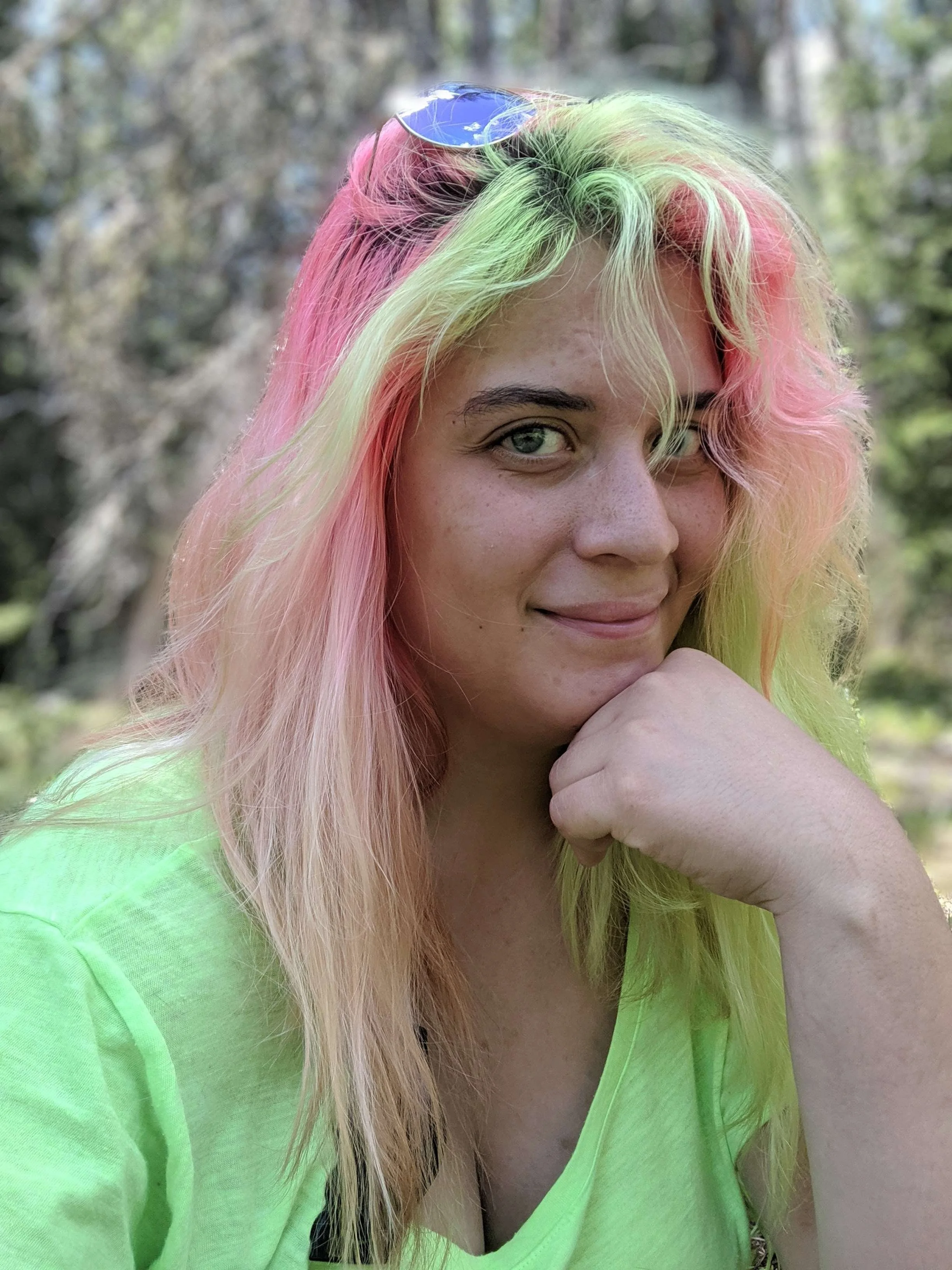 A woman with multi-colored hair in shades of pink, green, and yellow, wearing a neon green shirt and sunglasses on her head, smiling and looking at the camera outdoors.