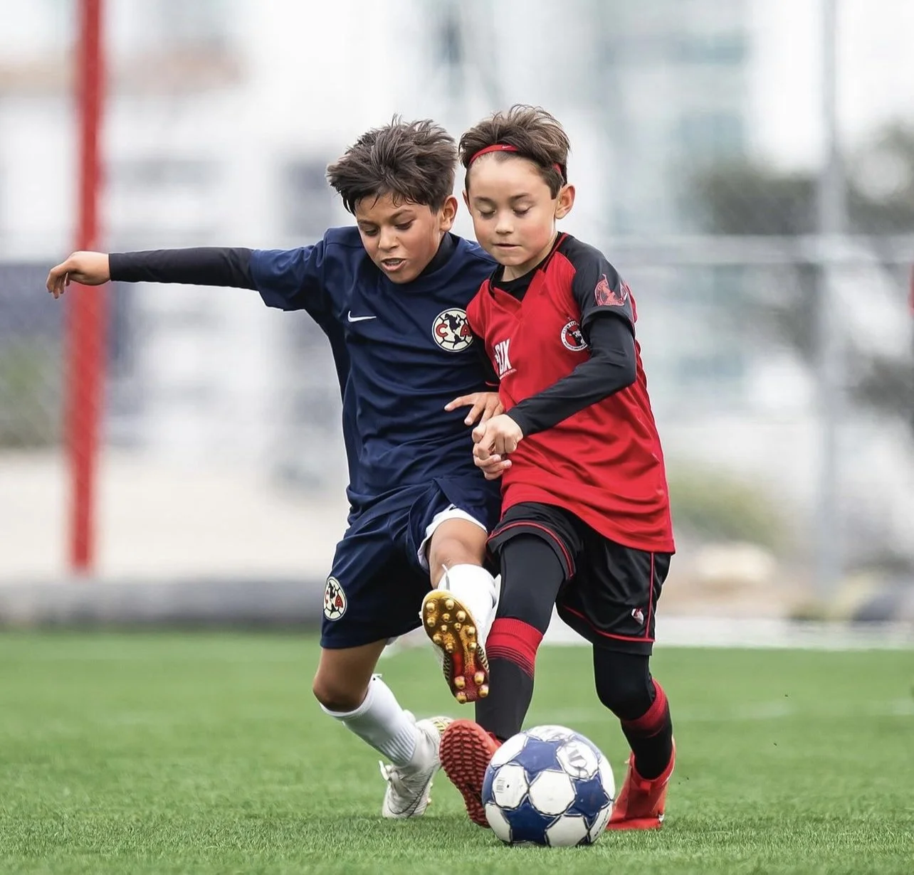Dos niños jugando fútbol, uno con uniforme azul y otro con uniforme rojo, ambos luchando por el balón en un campo de césped.
