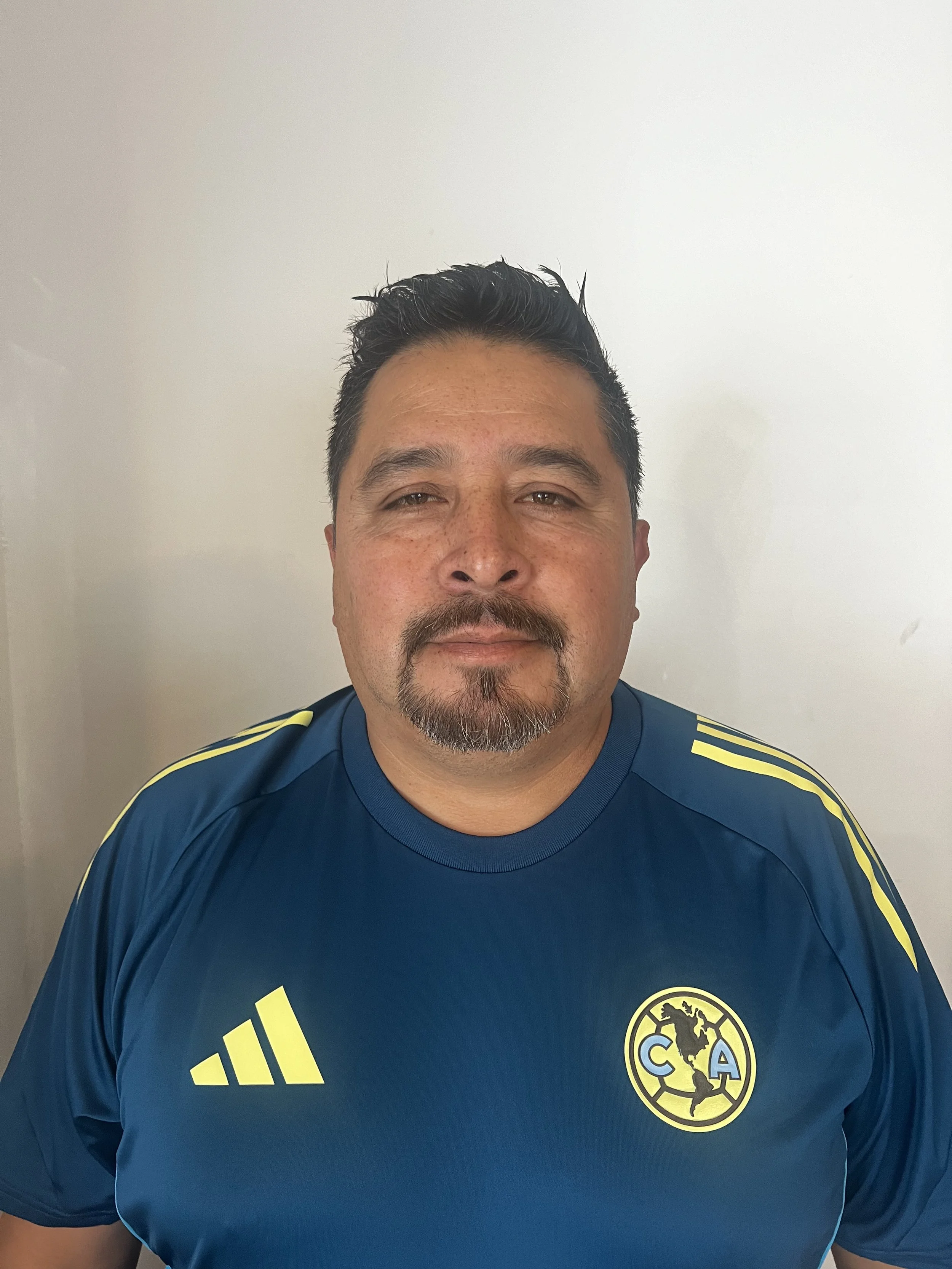 Hombre con barba y bigote, usando una camiseta azul de fútbol con rayas amarillas y un escudo del club América en el pecho.