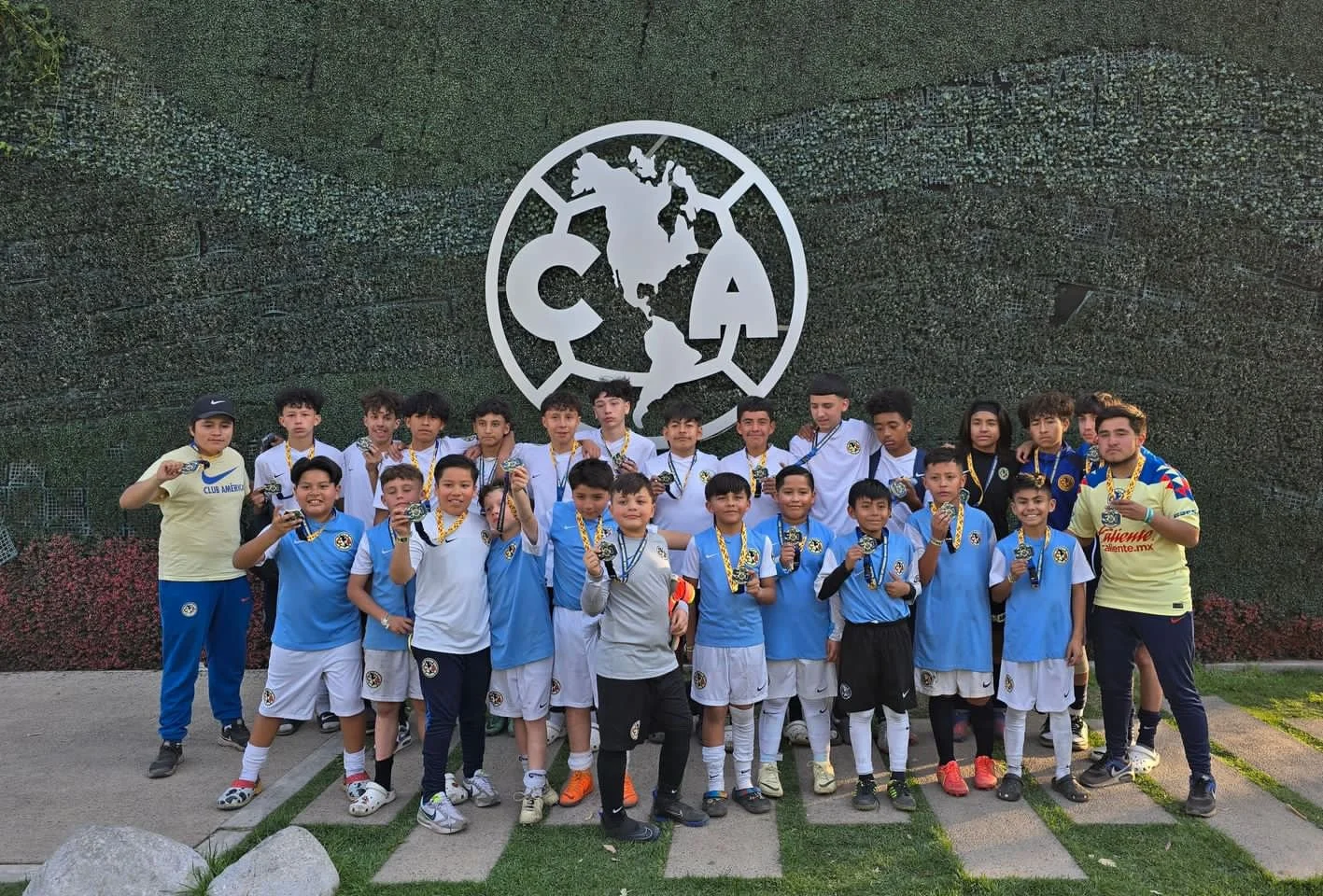 Grupo de niños y adolescentes con medallas posando frente a un logo de la FIFA en una pared de muro verde.