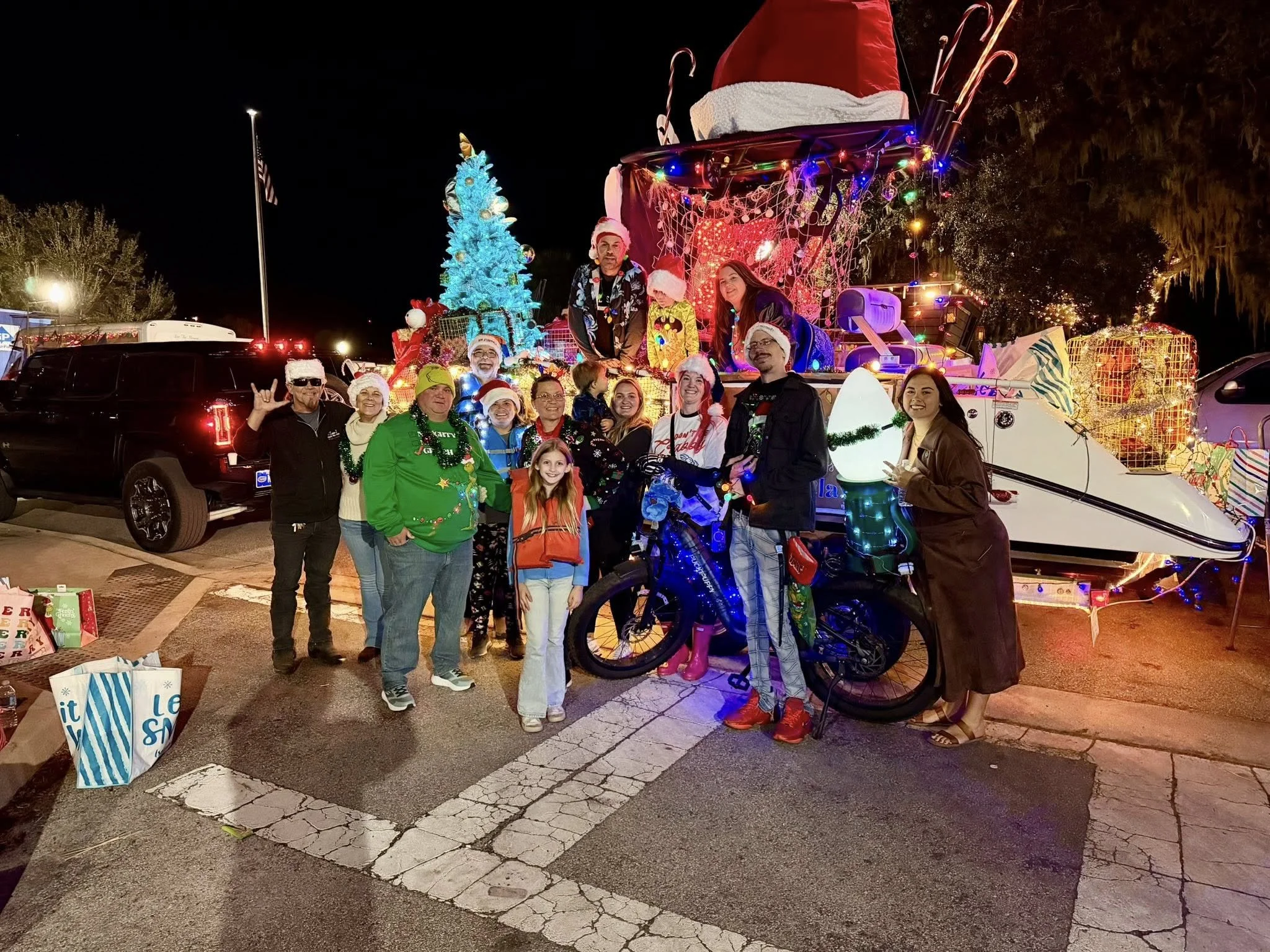 A group of people celebrating Christmas at night with a decorated float featuring a giant Santa hat on a sleigh, a Christmas tree, and wrapped presents. The group is smiling, wearing holiday sweaters and hats, with some making peace signs. There are cars and trees in the background.