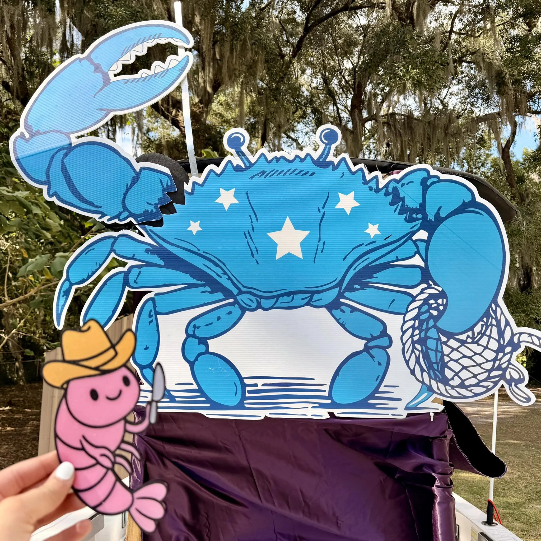 A colorful crab-shaped kite with stars on its back, held in front of a larger crab kite on display outdoors, with trees and sunlight in the background.
