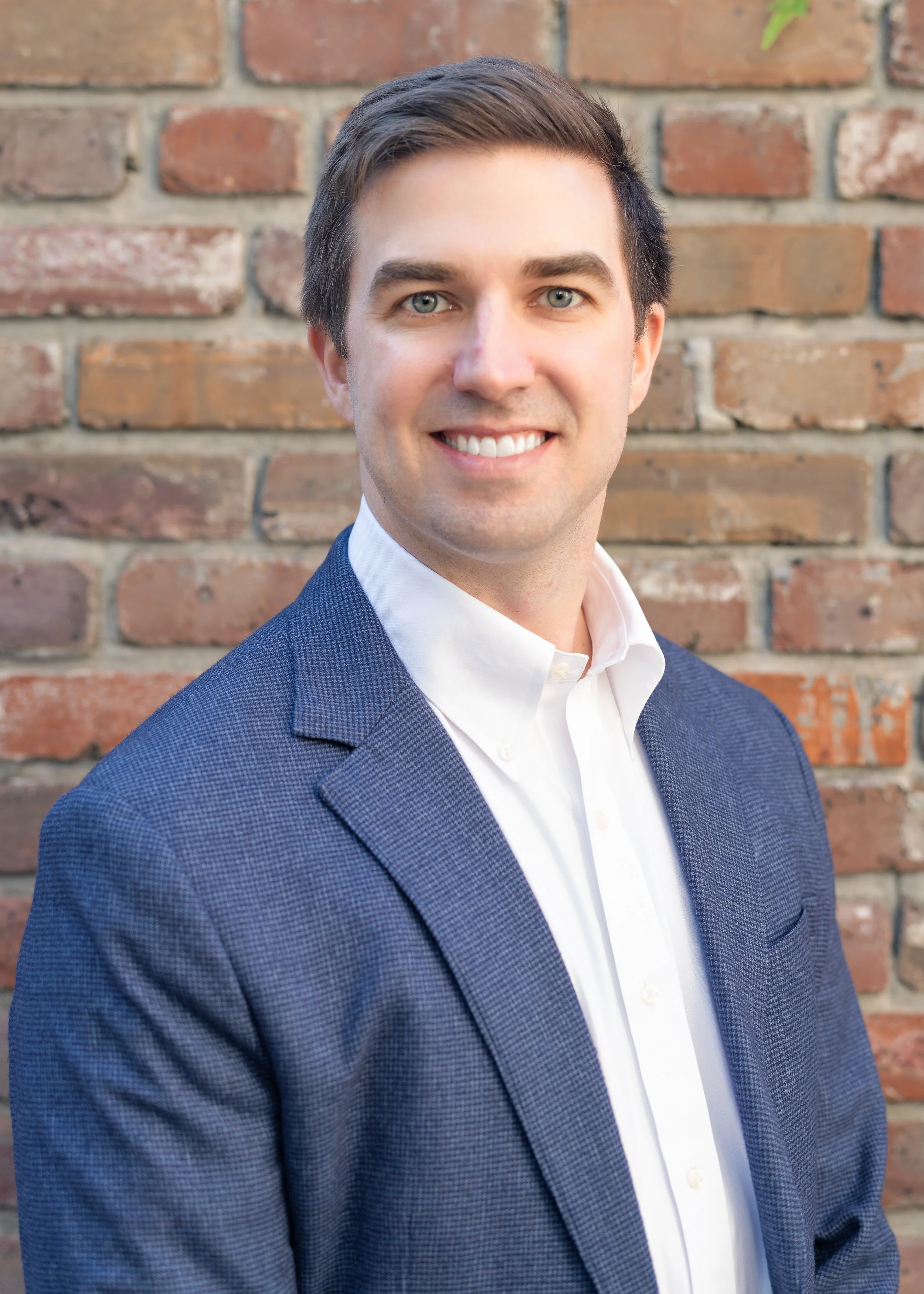 Portrait of a surgeon or doctor with light skin, blue eyes, short dark hair, smiling, wearing a white shirt and blue blazer, standing in front of a brick wall.