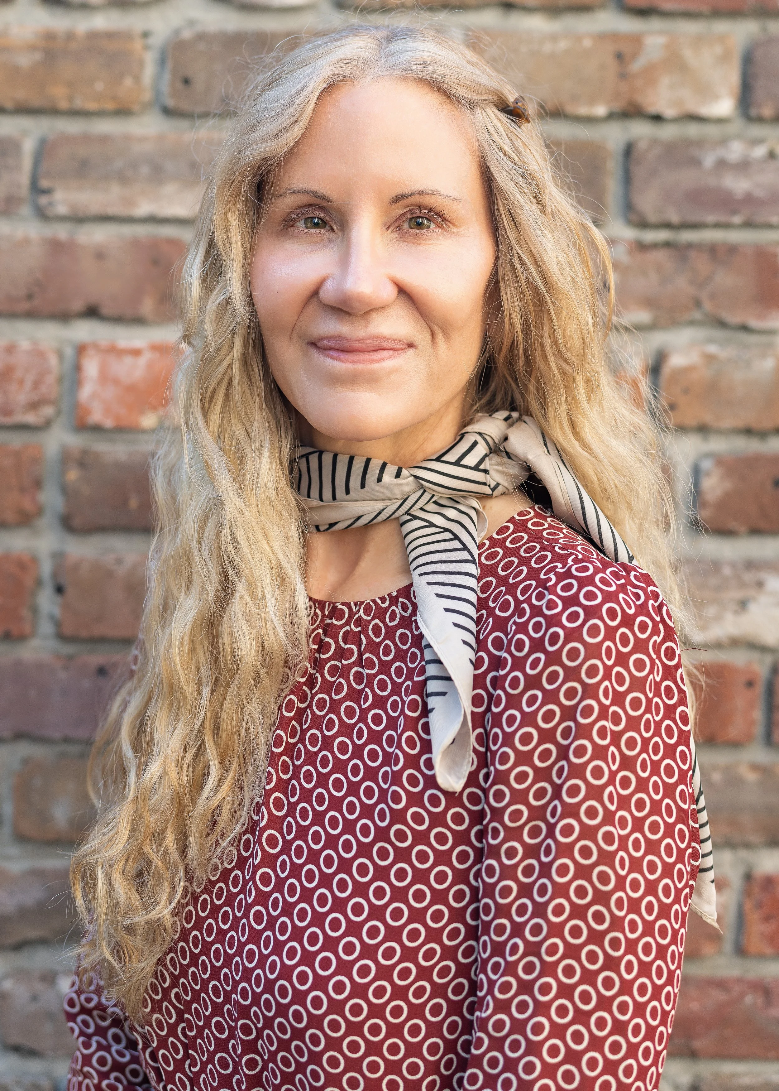 A surgeon or doctor with long curly blonde hair, wearing a red patterned top and a beige and black striped scarf, standing in front of a brick wall.