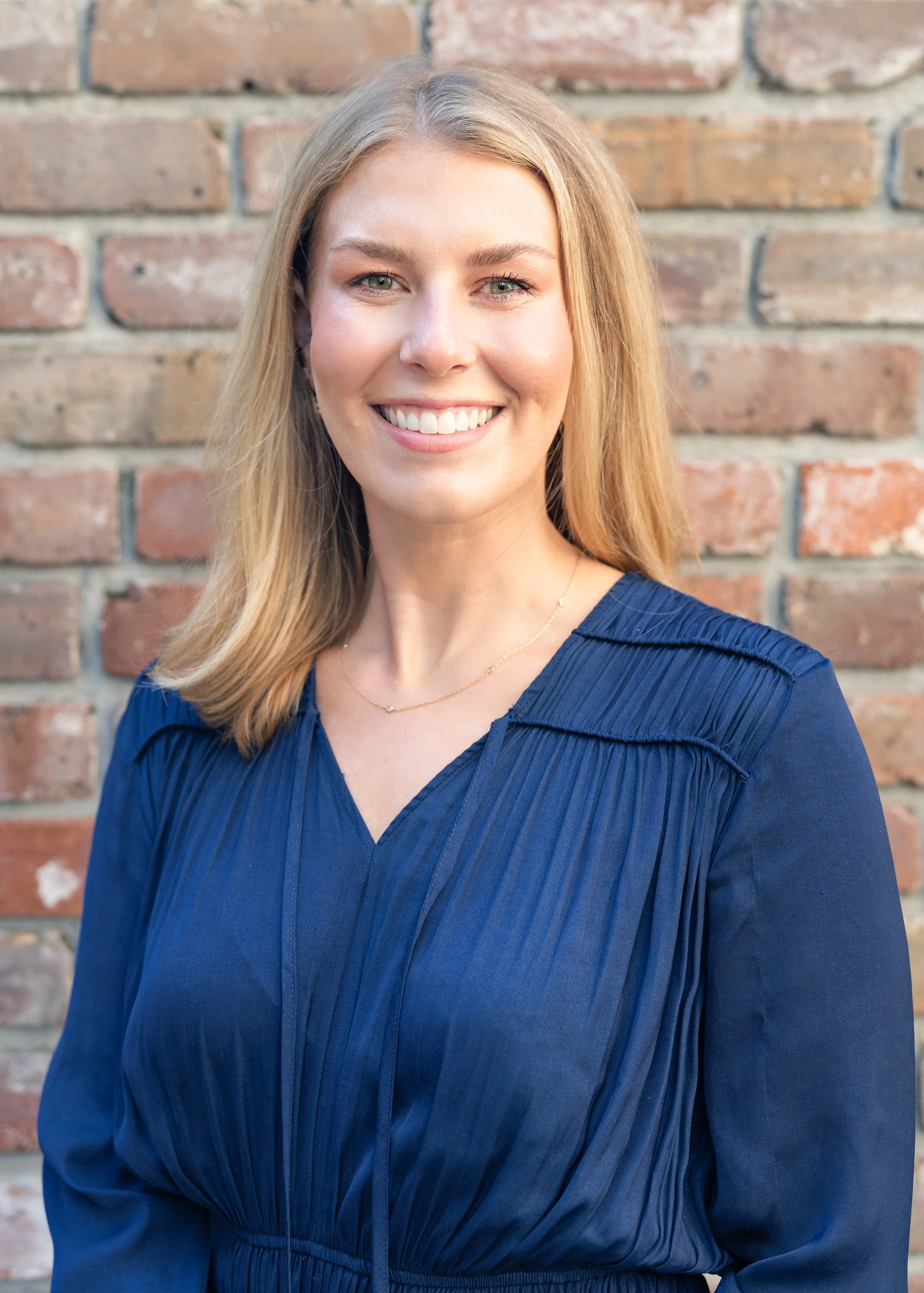 A woman with blonde hair and green eyes smiling in front of a brick wall, wearing a dark blue blouse and a delicate gold necklace.
