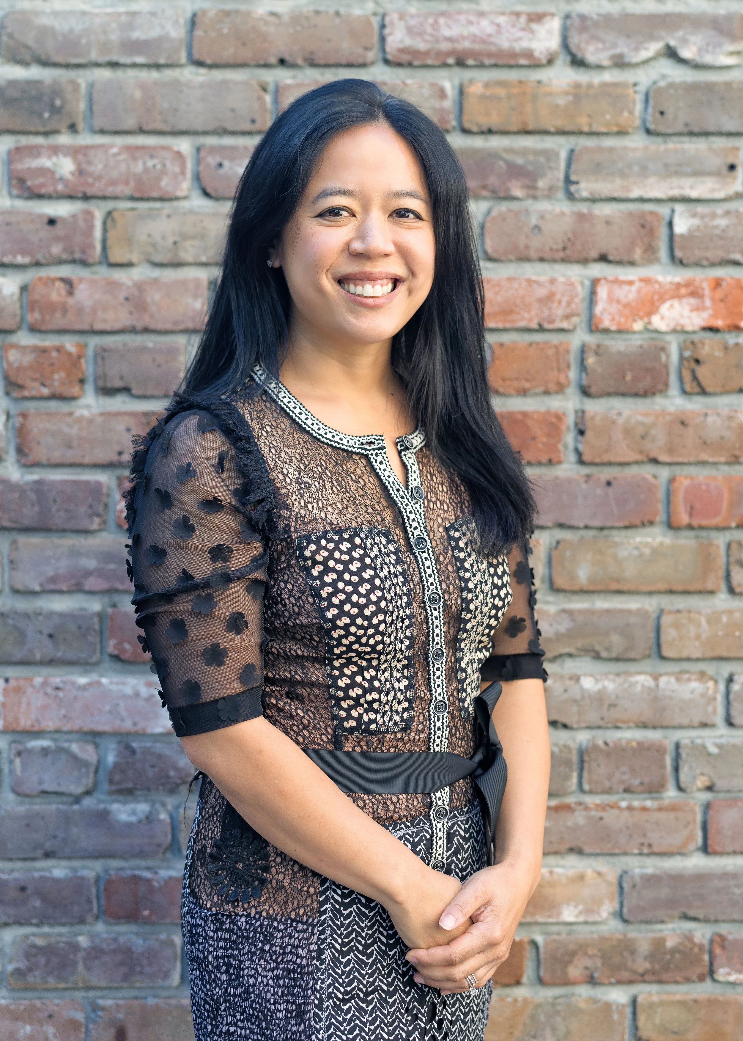 Smiling surgeon or doctor with black hair wearing a black and white patterned dress standing in front of a brick wall.