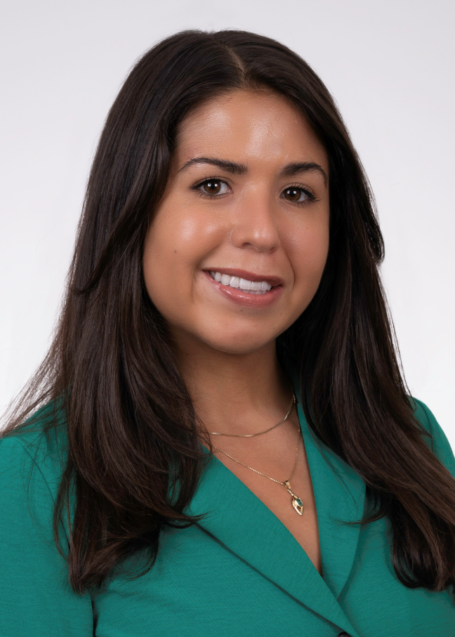 Close-up portrait of a young woman with long dark brown hair, smiling, wearing a green blazer and layered gold necklaces, against a plain white background.
