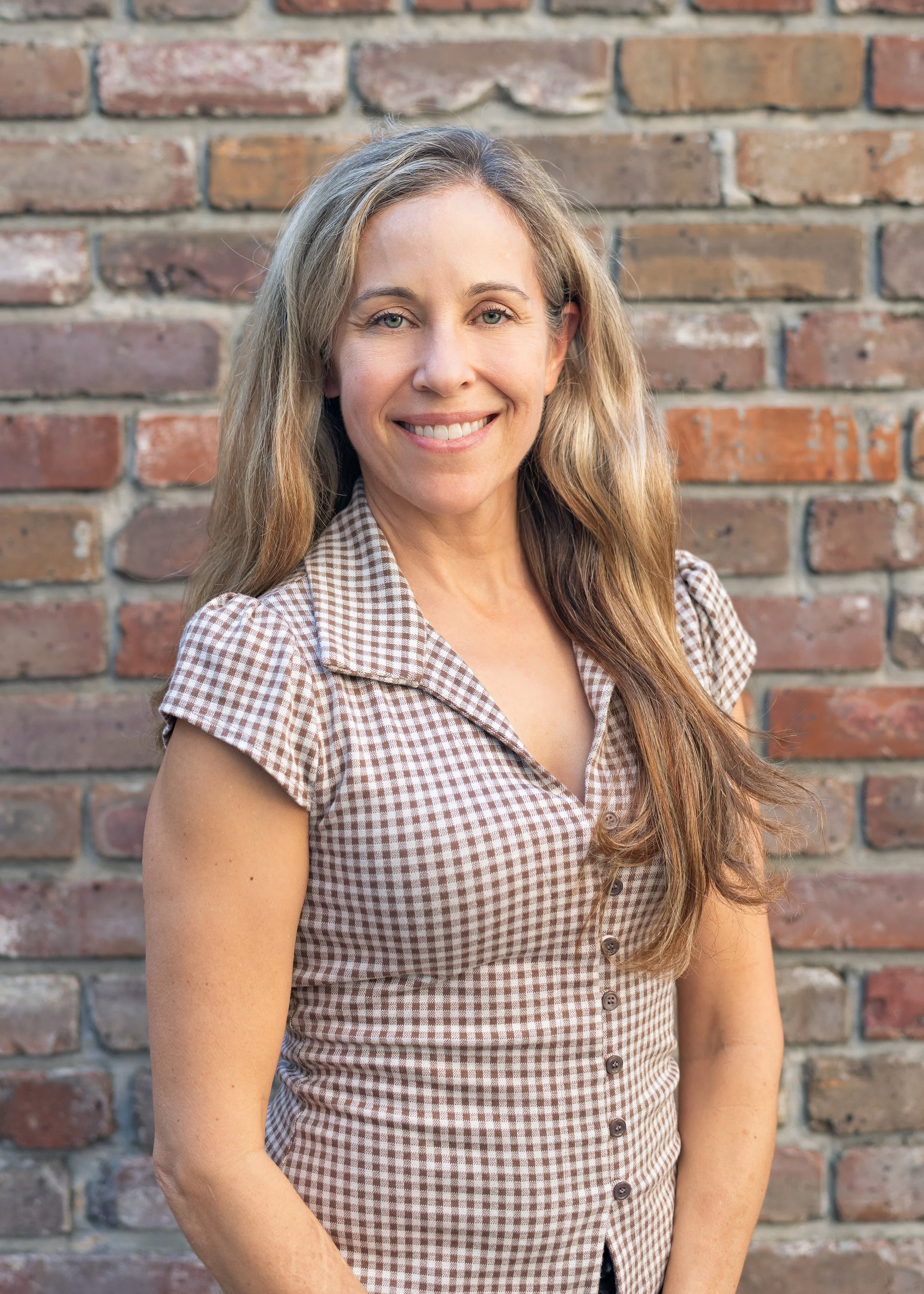 A surgeon or doctor with long, wavy blonde hair smiling and standing in front of a brick wall, wearing a short-sleeved, button-up, brown and white checkered dress.