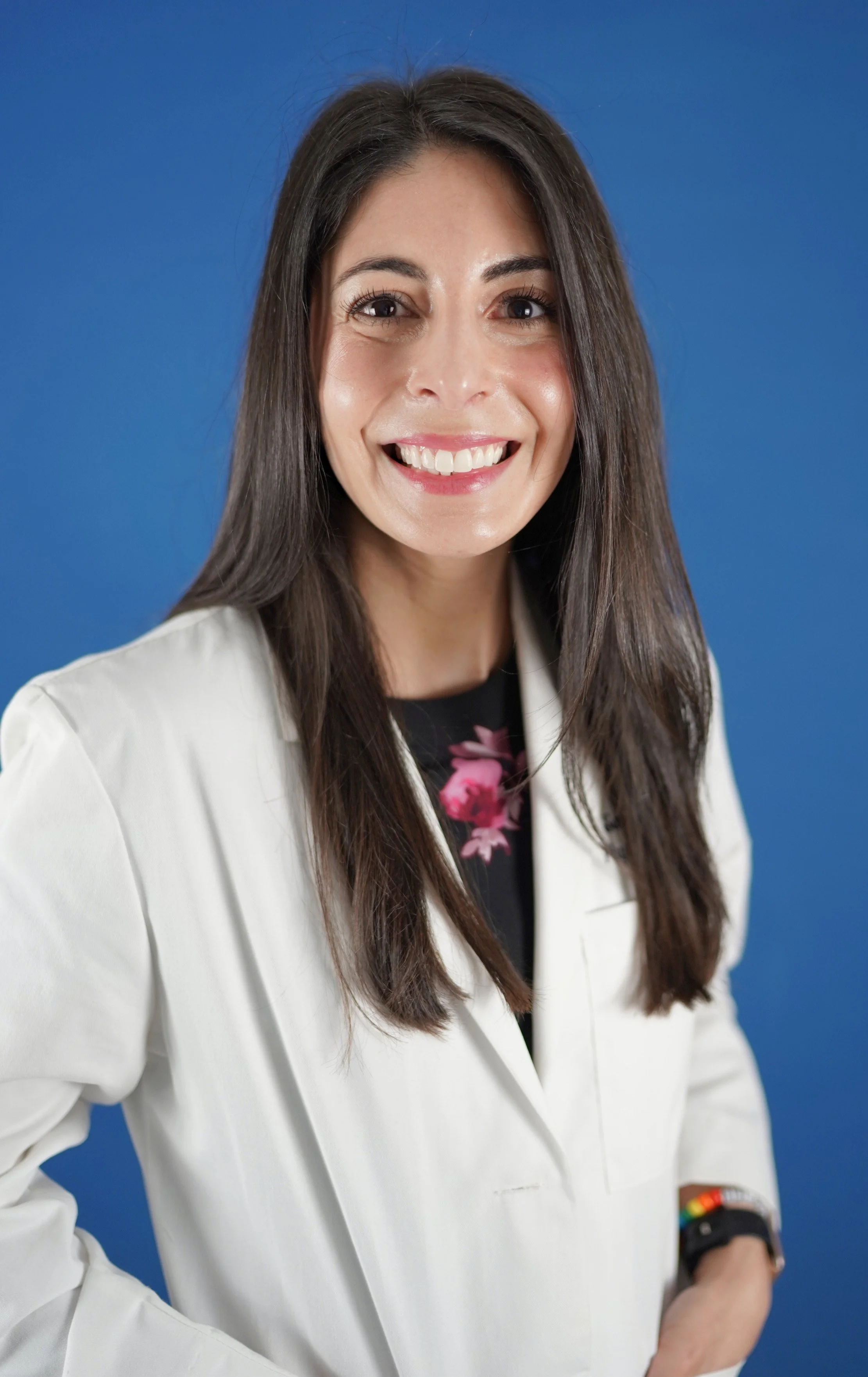 Smiling woman with long dark hair, wearing a white blazer over a black top with pink floral design, standing against a solid blue background.