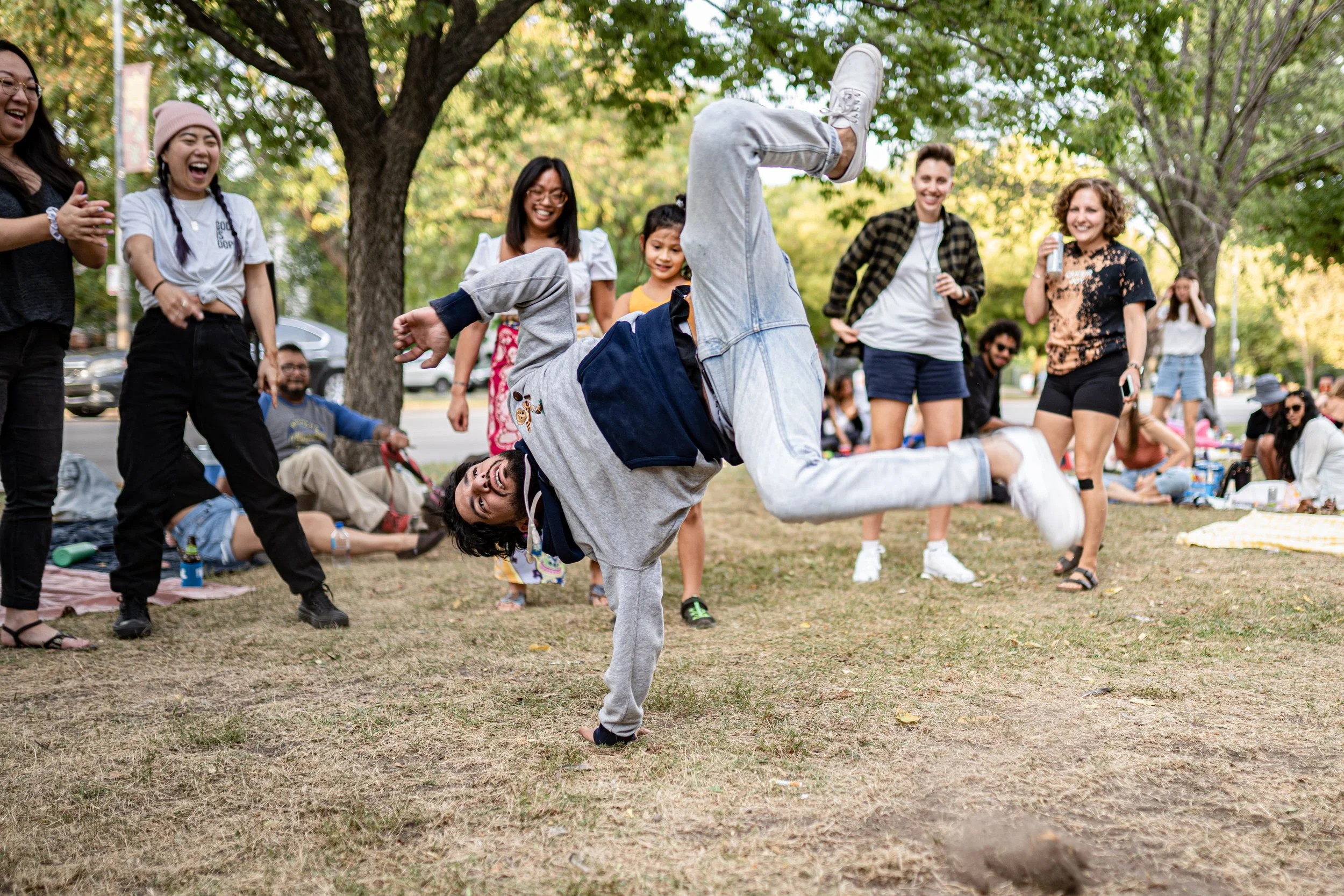 Group of people outdoors watching a man performing a breakdance move on the grass in a park with trees, while others laugh and enjoy the event.