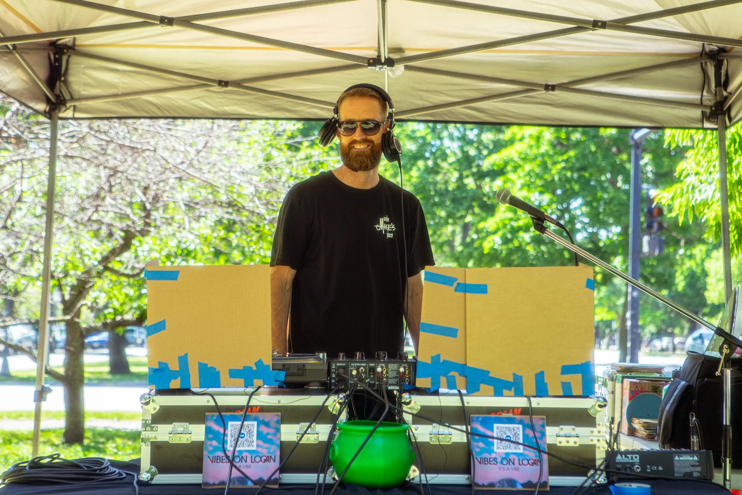 A man with a beard, sunglasses, headphones, and a black t-shirt standing behind a DJ setup under a canopy at an outdoor event, with trees and greenery in the background.
