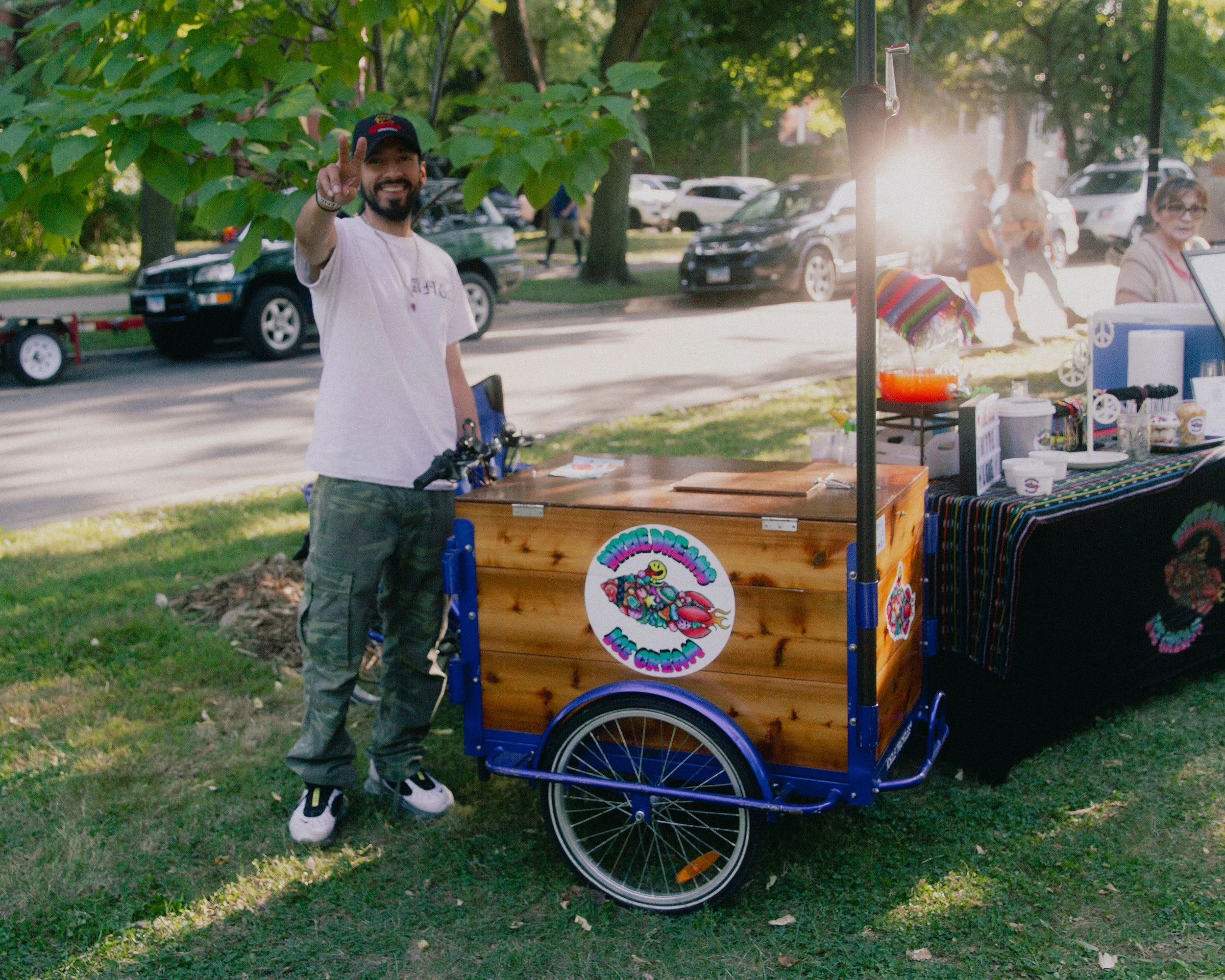 A man with a beard, wearing a black cap, white t-shirt, and green pants, stands beside a colorful, decorated ice cream or dessert cart, making a peace sign with his right hand at an outdoor event in a park with trees and parked cars in the background.
