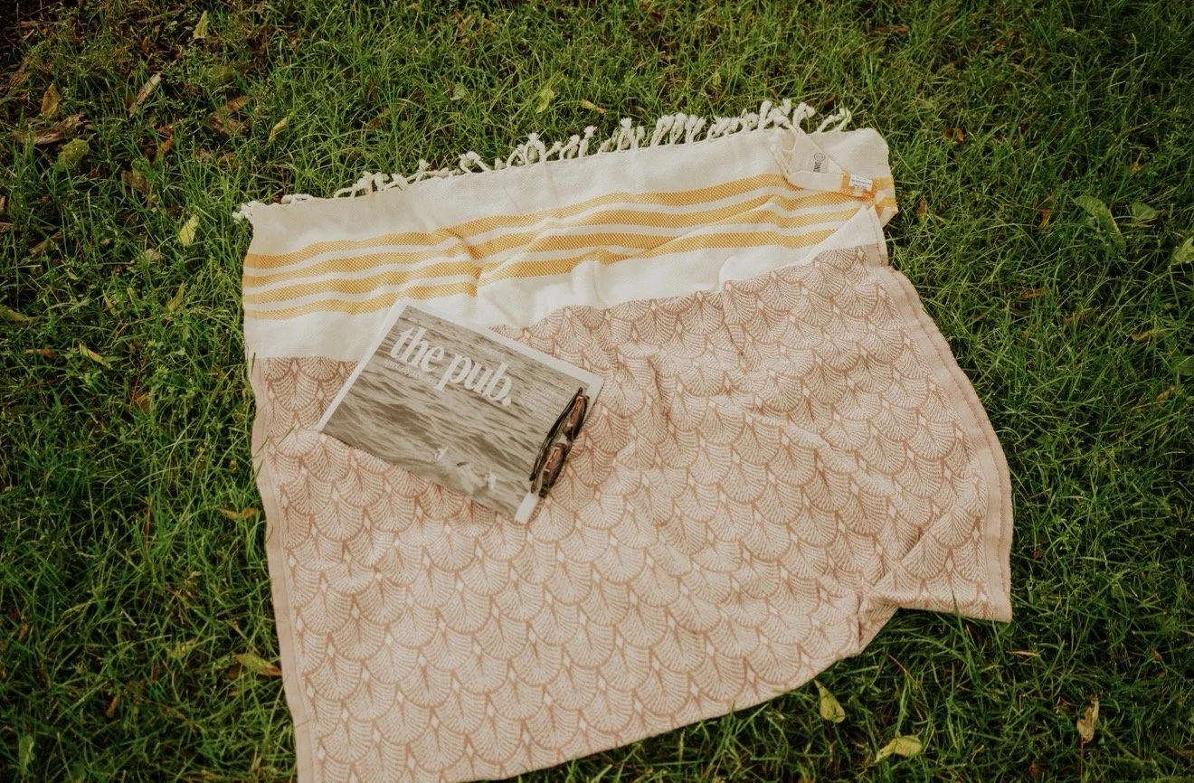 A striped beach towel with a lace pattern, laid on grass, with a folded newspaper and a pair of eyeglasses on top.