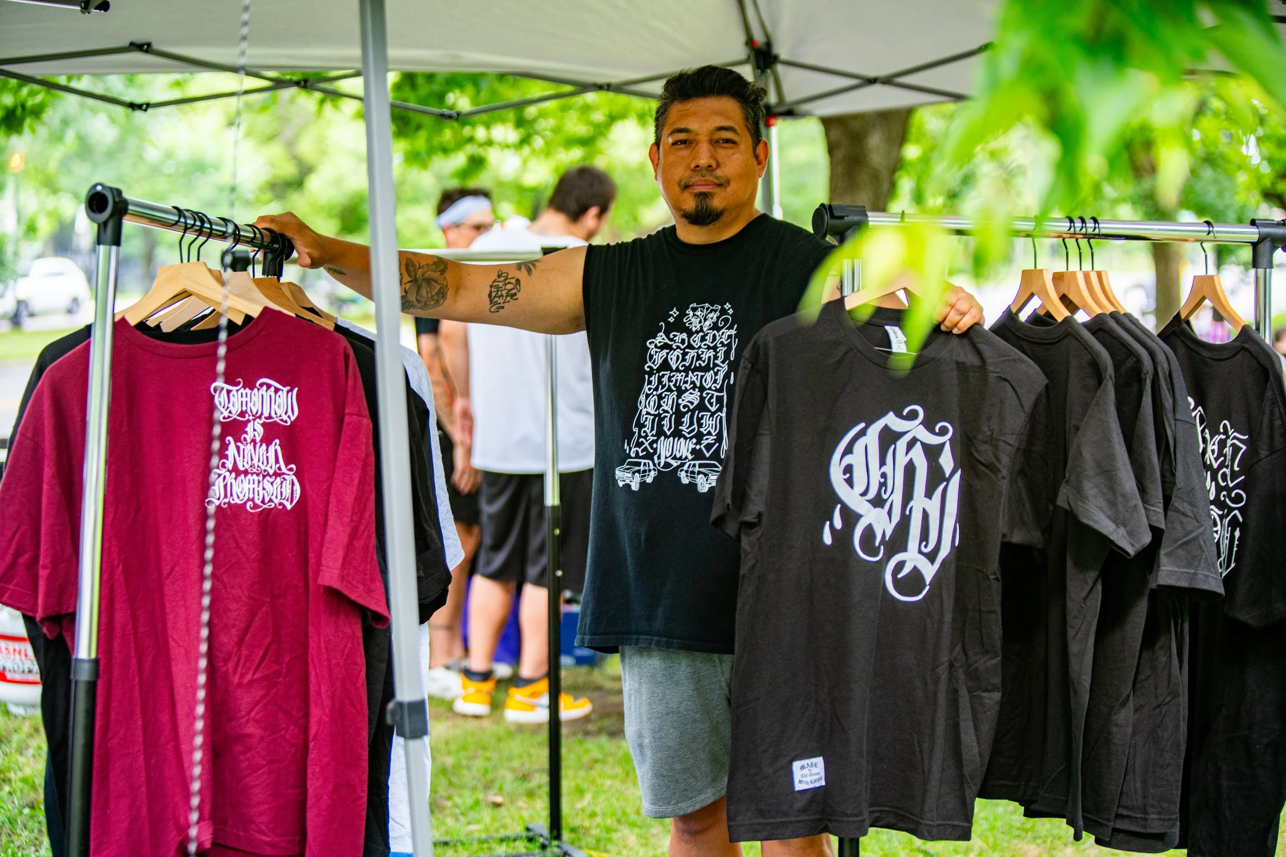 Man standing at an outdoor clothing market booth with black and red T-shirts hanging on a rack under a white canopy, green trees in the background, and other people in the distance.