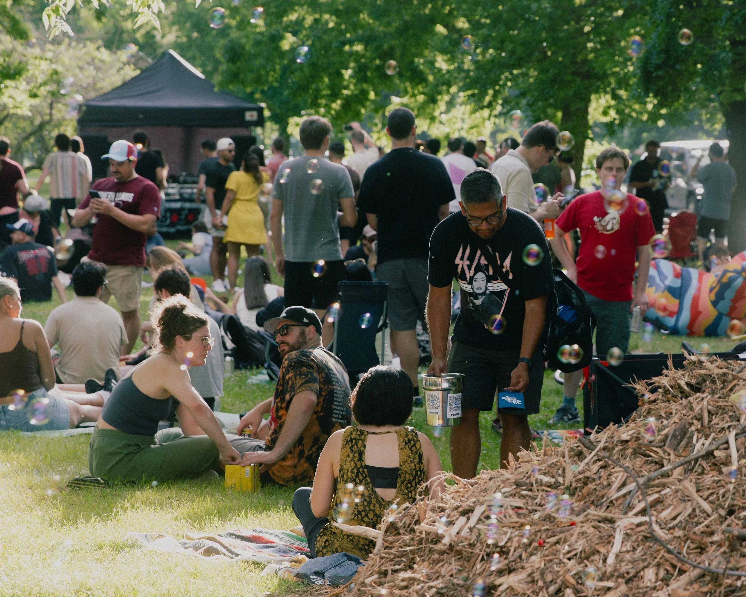 A group of people gathered in a park during daytime for an outdoor event, with some sitting on the grass and others standing, surrounded by trees and soap bubbles floating in the air.