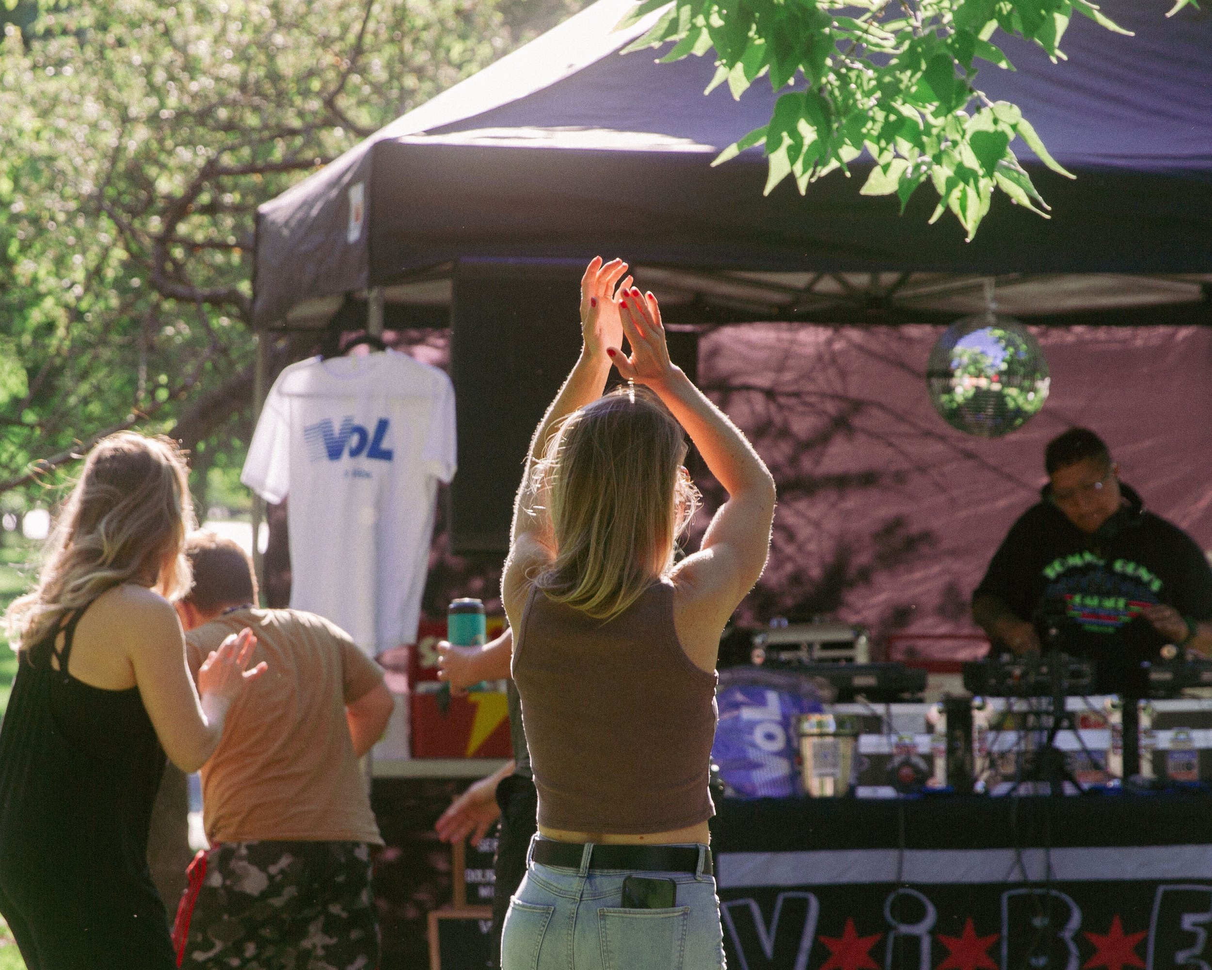 People outdoors, some with hands raised, near booths or stalls, green trees and a canopy overhead, sunlight filtering through, suggesting a public event or festival.