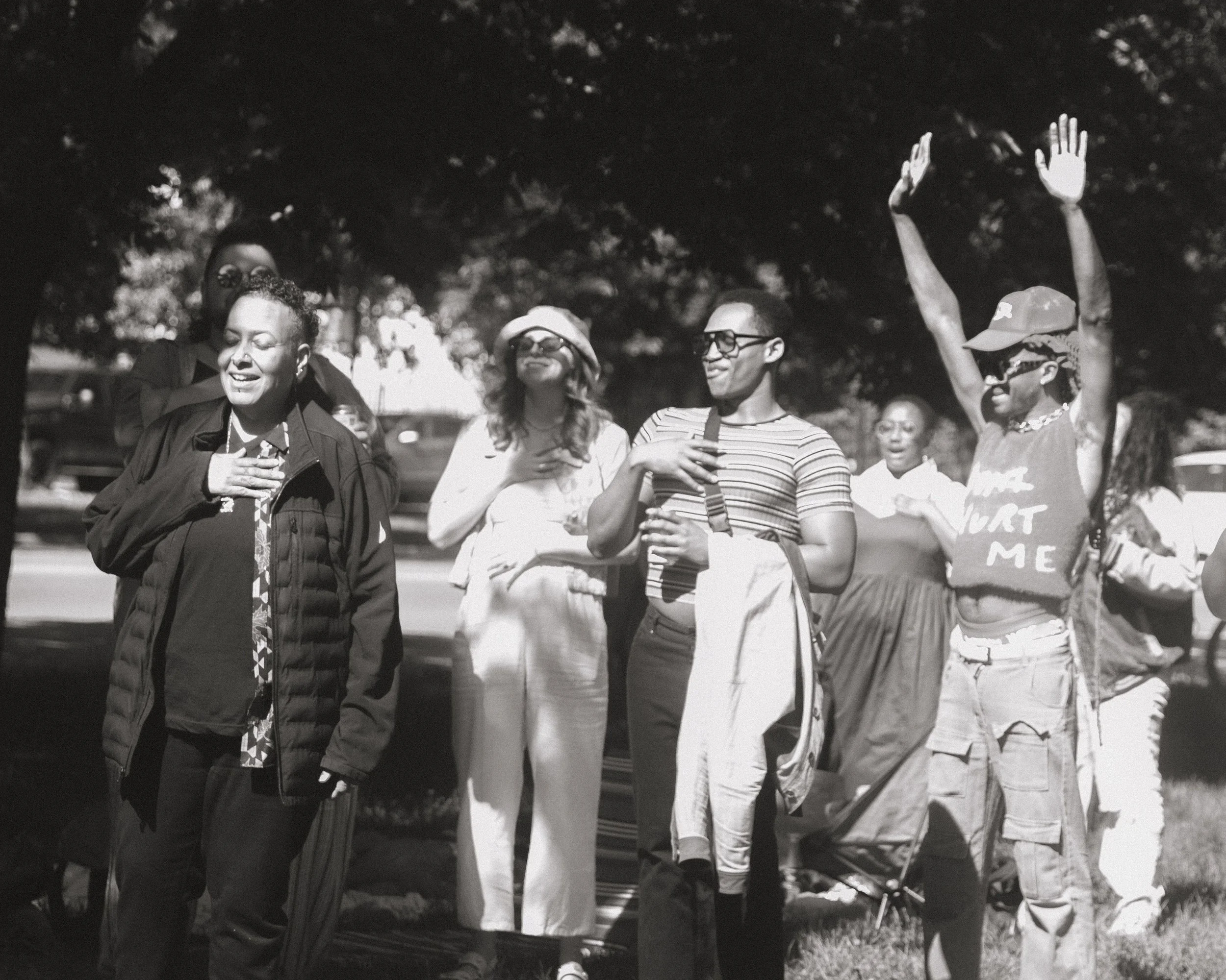 Group of people outdoors, walking and talking, some with sunglasses and casual clothing, under trees.