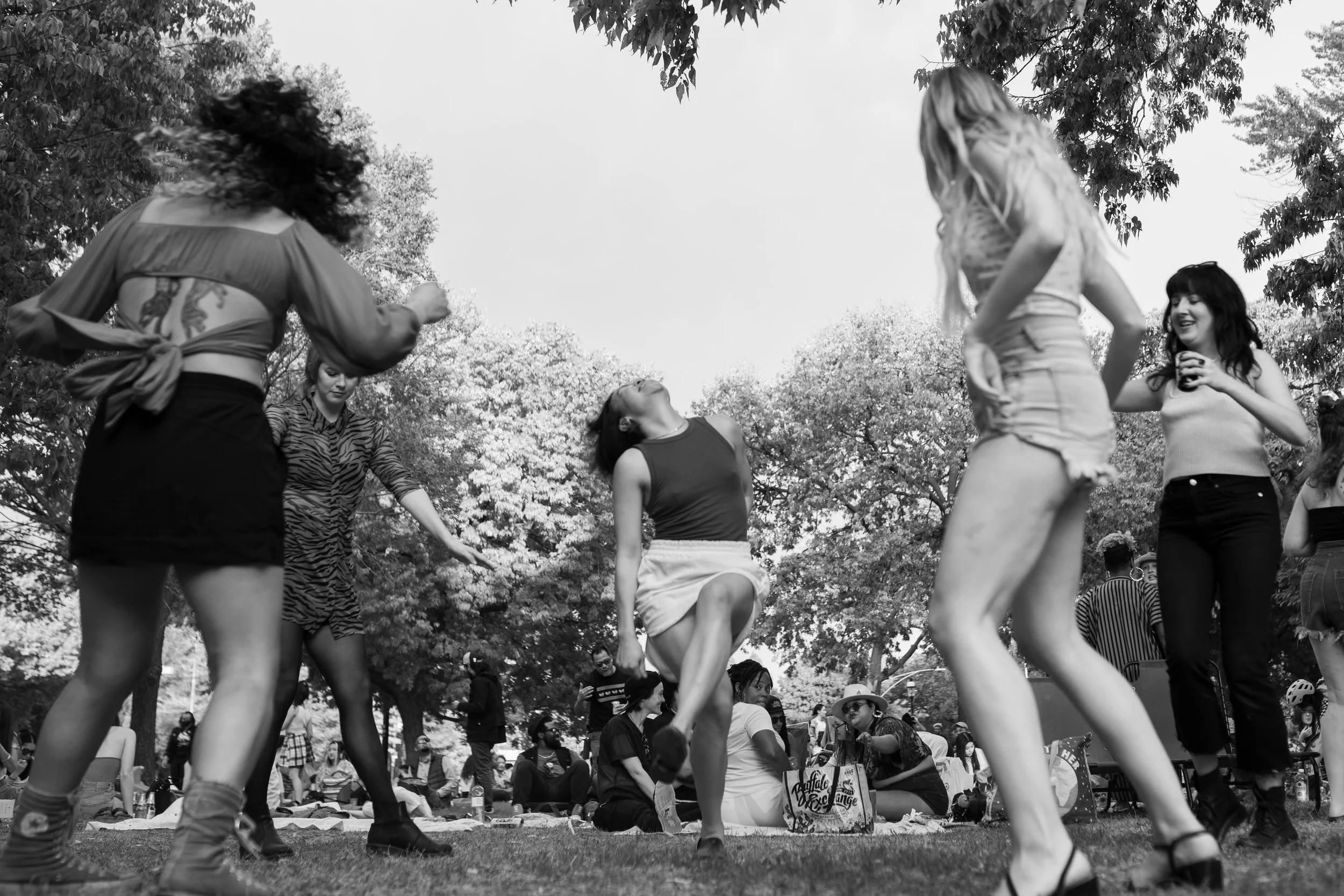 People dancing and playing in a park during an outdoor event with trees and spectators in the background.
