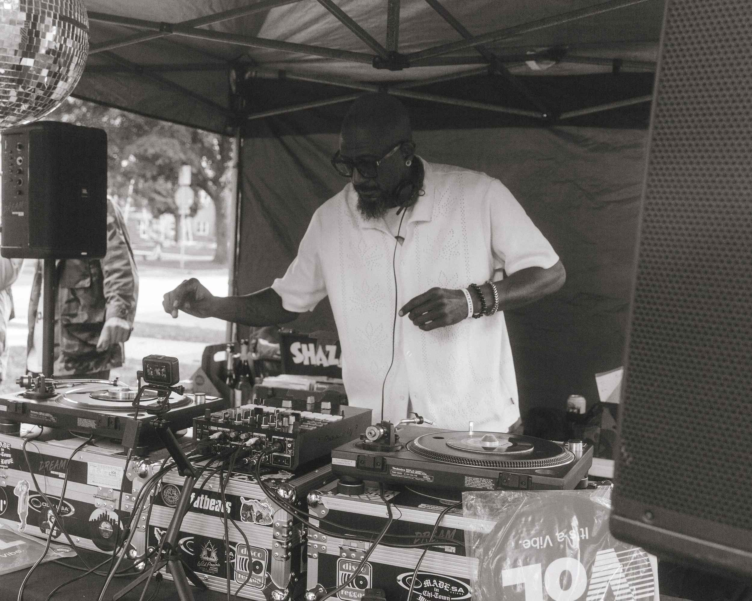 A DJ wearing glasses and headphones is performing at an outdoor event under a canopy. DJ equipment, including turntables and a mixer, are on the table in front. A disco ball is hanging from the canopy in the top left corner of the image.