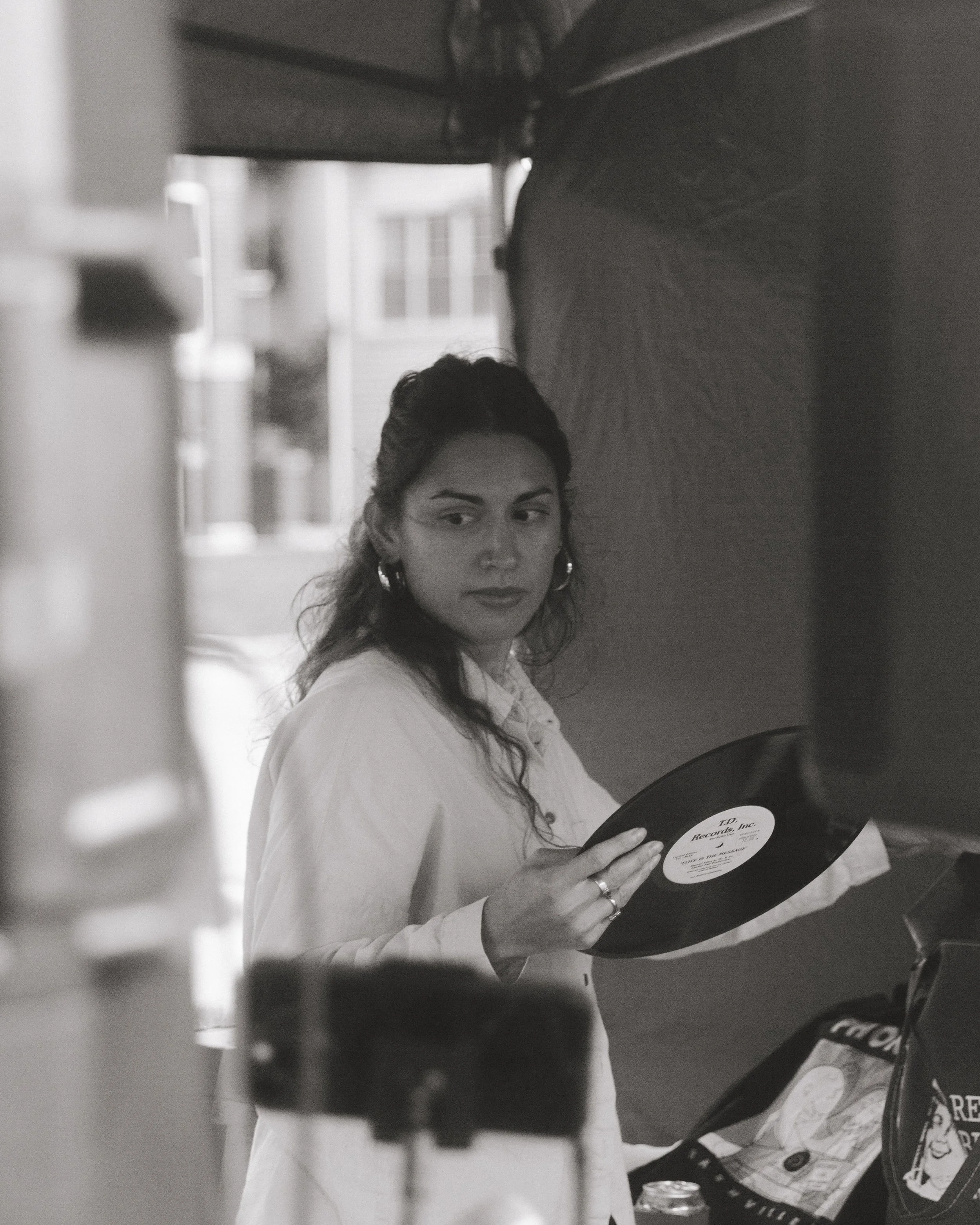 A woman with dark hair and earrings, wearing a white blouse, looking directly at the camera while holding a vinyl record in a room with a window in the background.
