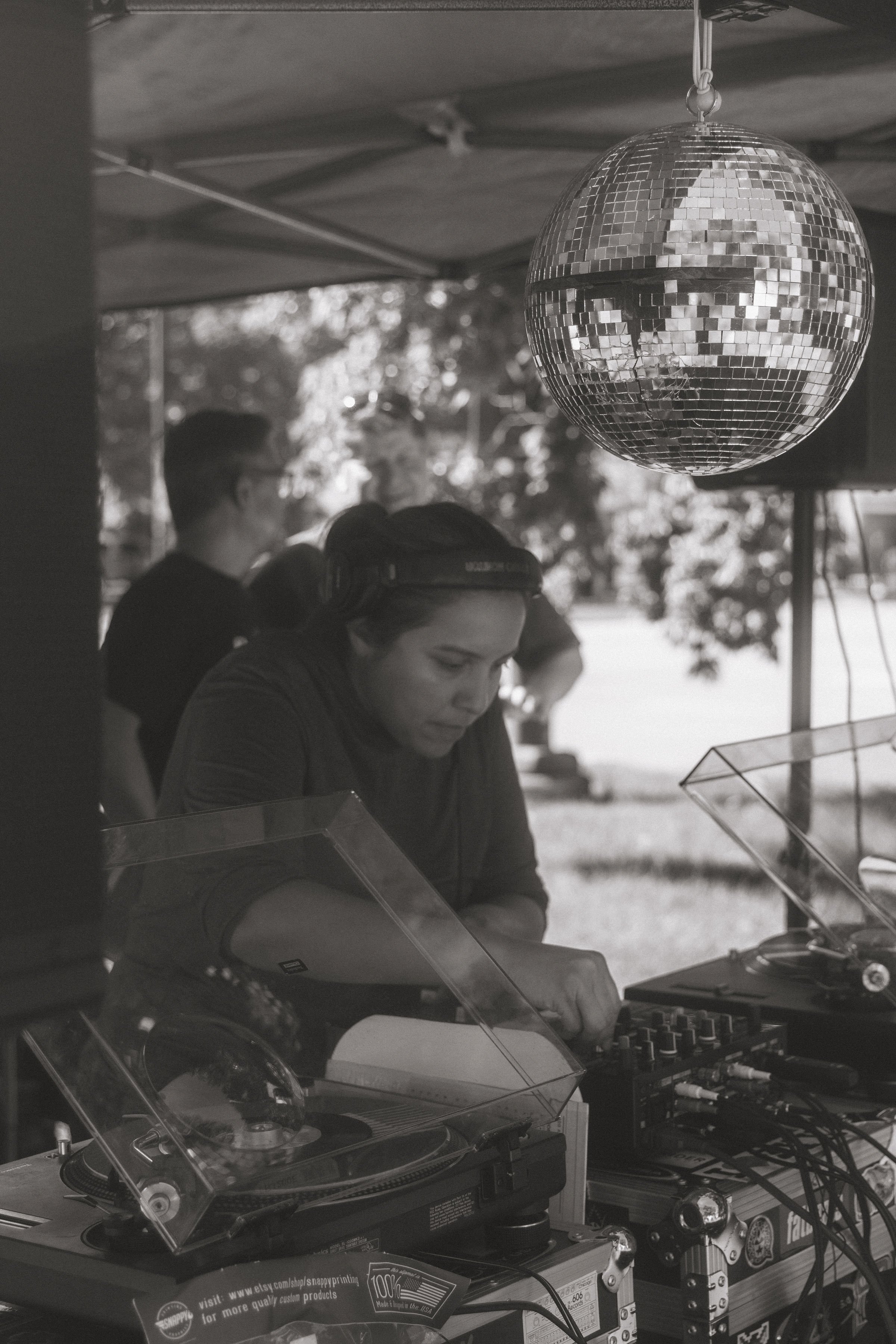 A black and white photo of several people, including a woman with headphones, operating DJ equipment outdoors under a canopy. A large disco ball hangs from the ceiling.