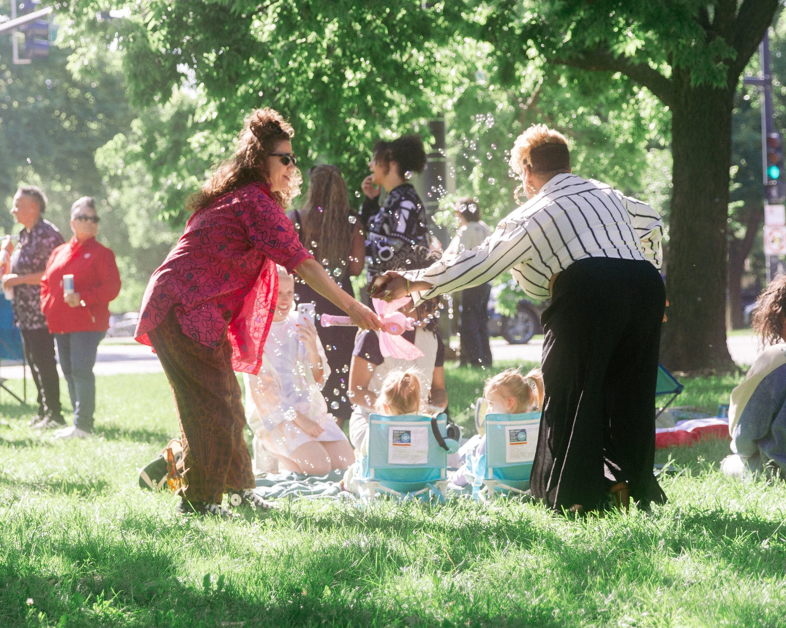 People enjoying a sunny outdoor gathering in a park, with women and children sitting on the grass and engaging with each other, under green trees.