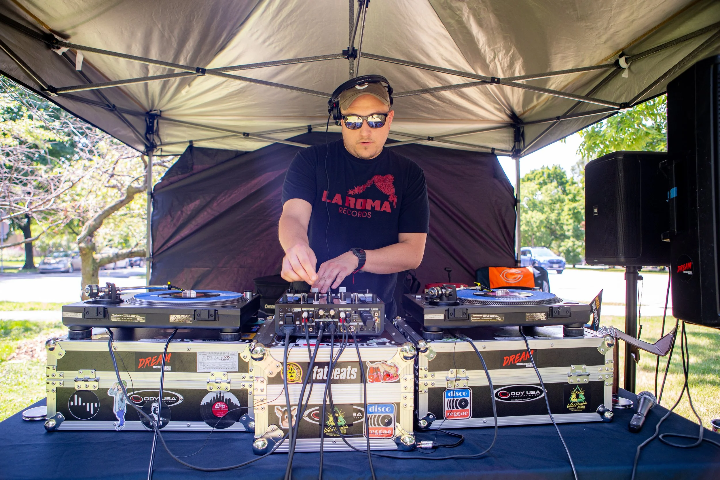 A DJ wearing sunglasses, a cap, and headphones performing under a canopy at an outdoor event, with trees and parked cars in the background.