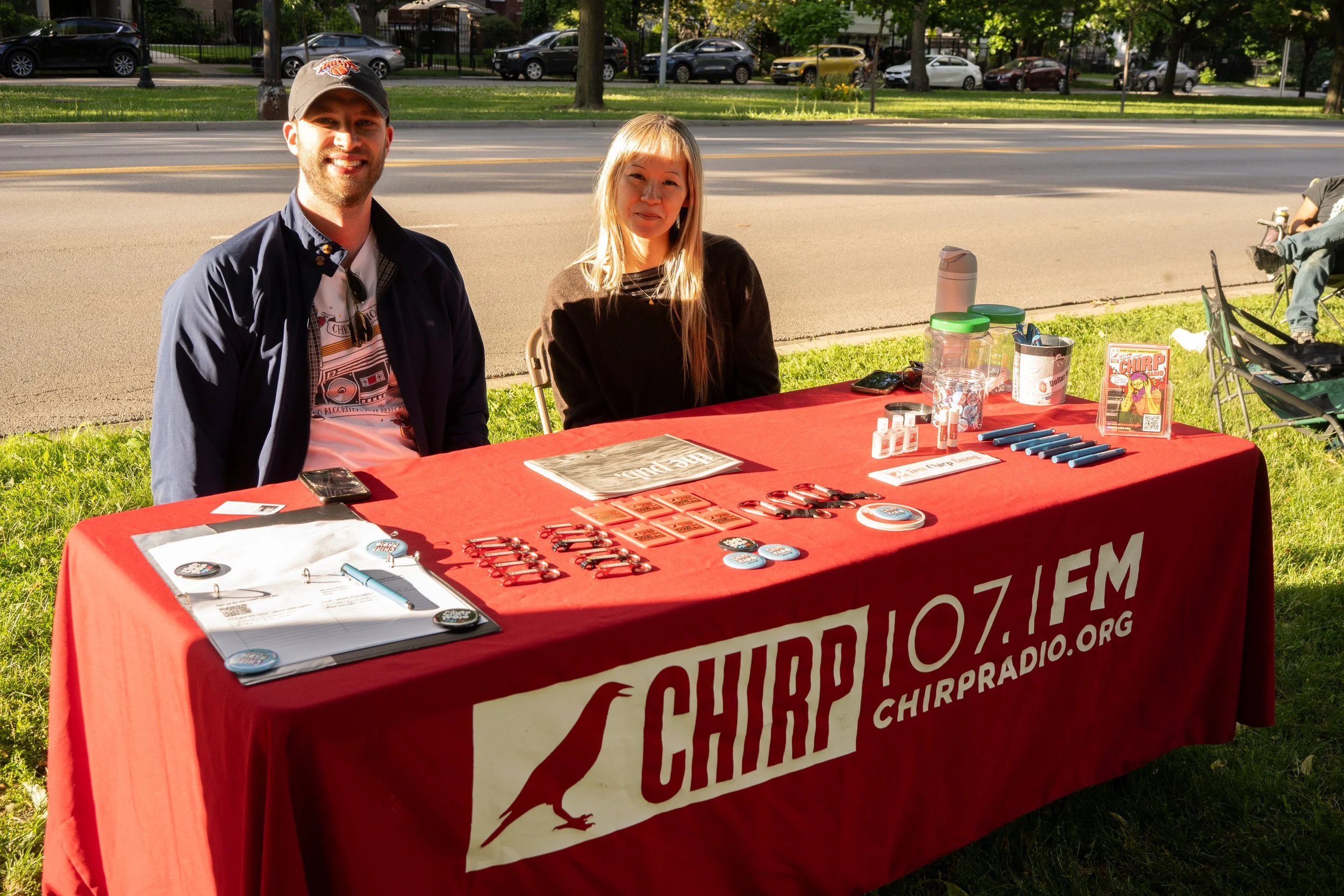 Two people sitting at a table outdoors with promotional materials, buttons, and pens, behind a red tablecloth promoting a radio station, with parked cars and trees in the background.