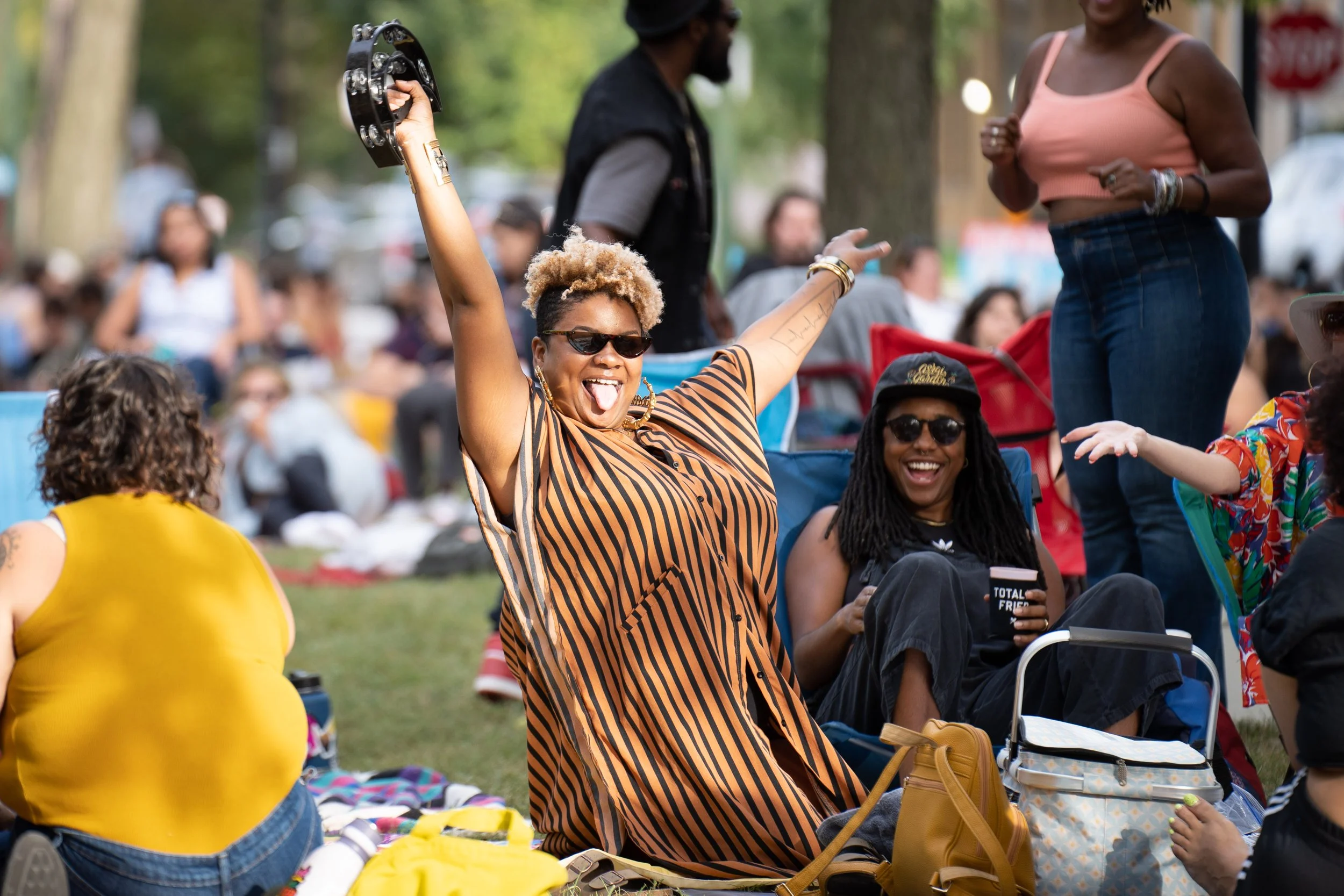 People enjoying outdoor gathering, smiling, sitting on the grass, with one woman dancing holding a tambourine, others sitting and chatting in a park setting.