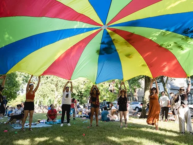 Group of women holding a large colorful beach umbrella outdoors in a park with trees and people sitting on a blanket.
