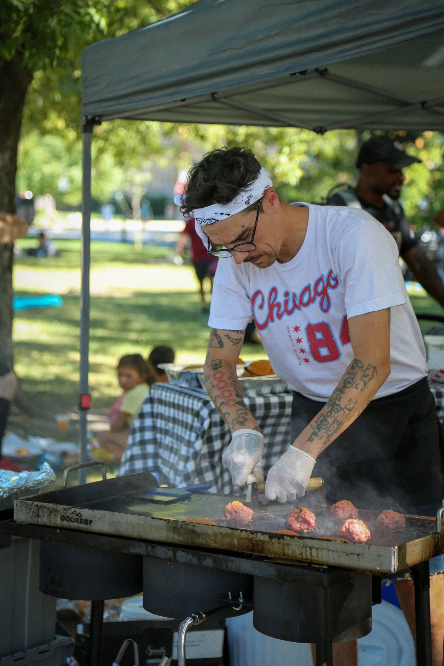 A man with tattoos and glasses wearing a white Chicago baseball t-shirt, a bandana, and gloves cooking burger patties on a flat-top grill at an outdoor event, with trees and other people visible in the background.