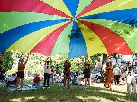 People holding a large, colorful rainbow umbrella in a park during the day.