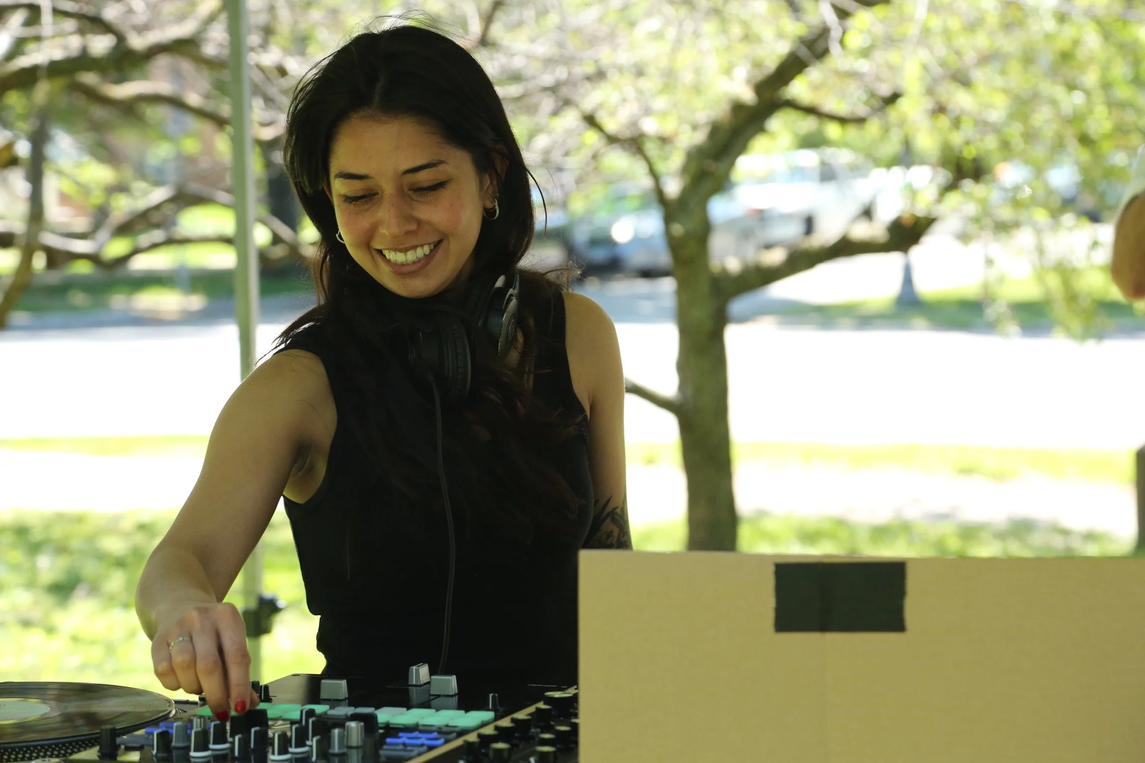 A woman with dark hair and earrings smiling while DJing outdoors with trees and sunlight in the background.