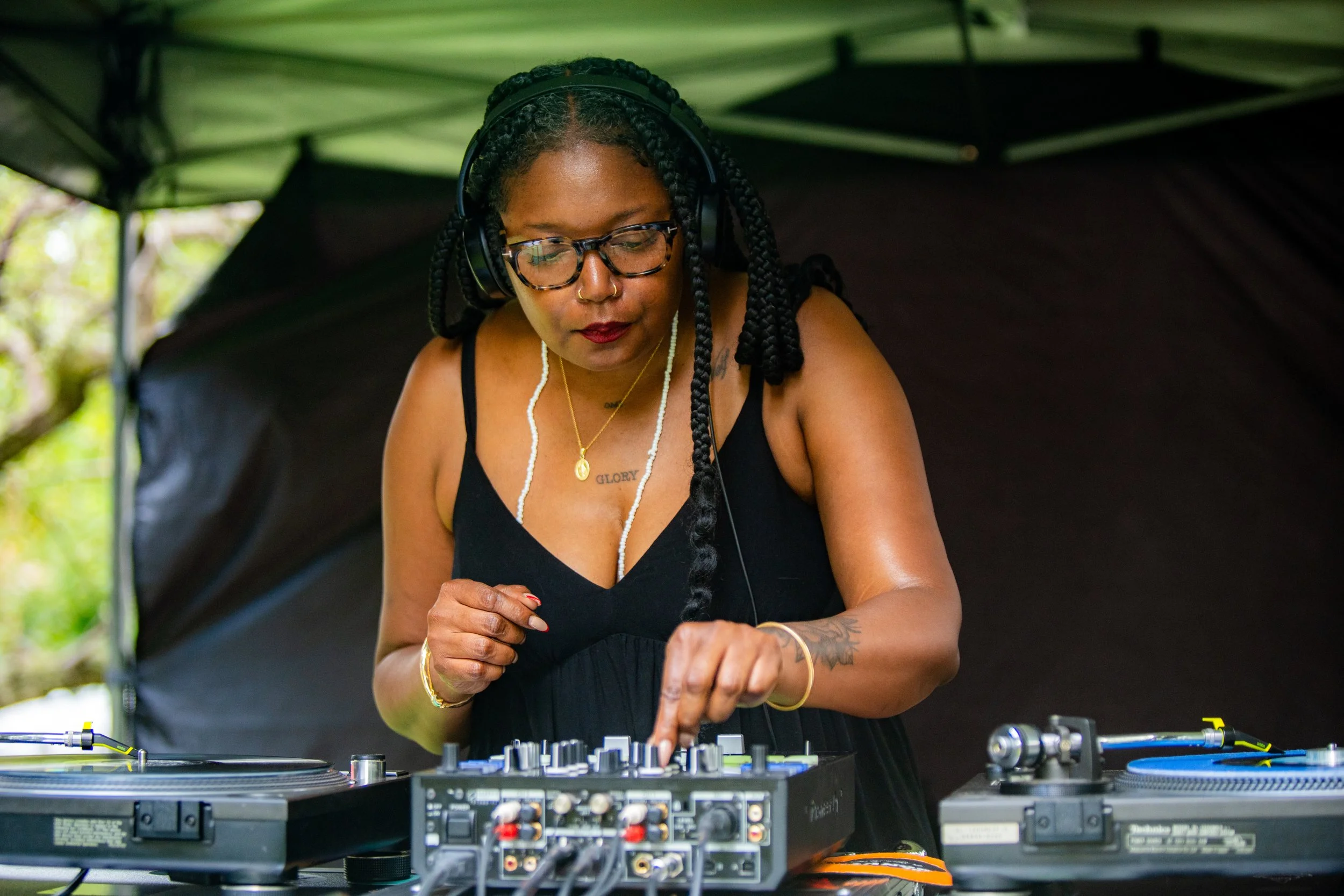 A female DJ with braided hair, glasses, and tattoos, wearing headphones, operating DJ equipment outdoors under a canopy.
