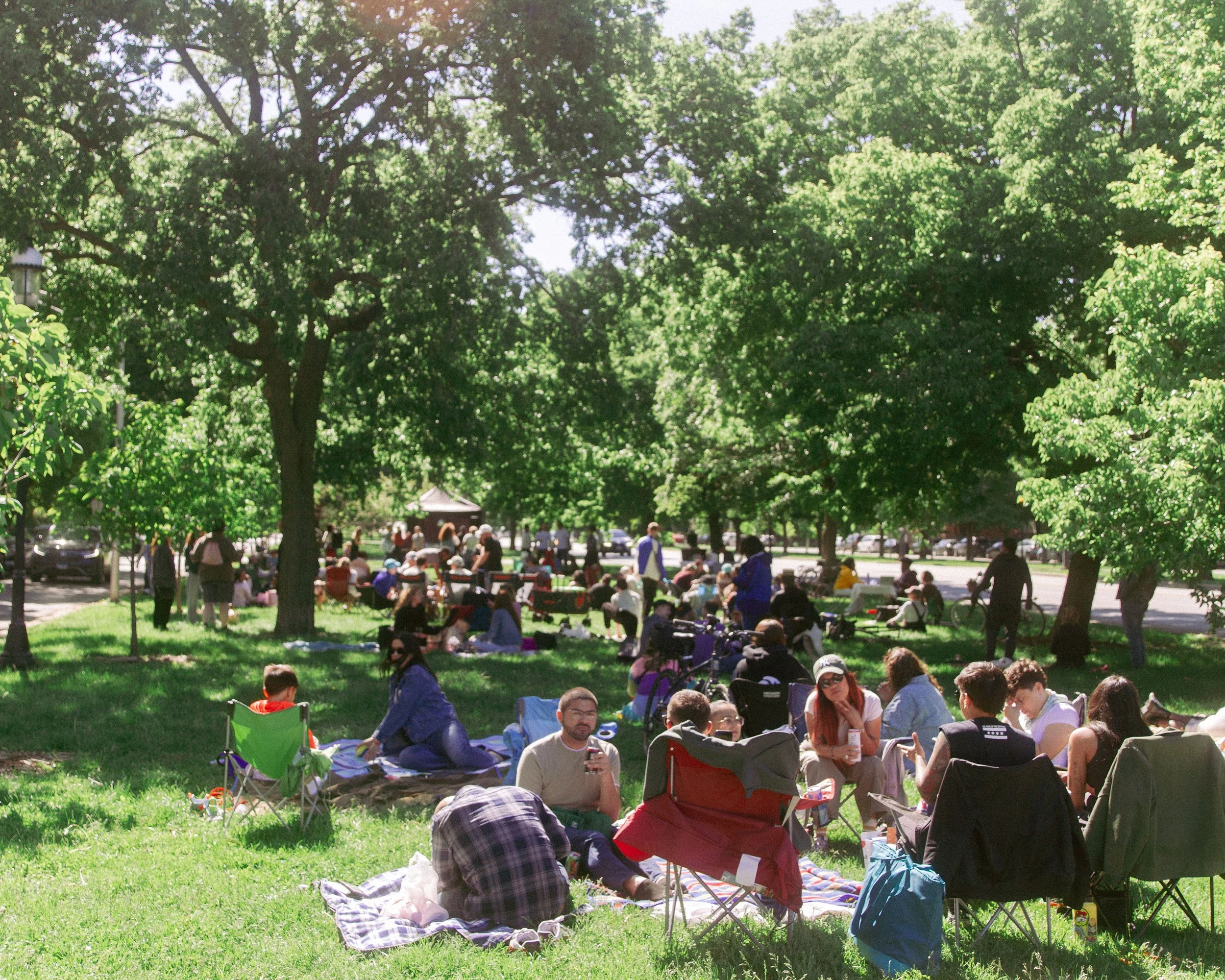 A park filled with people sitting on blankets and lawn chairs under large green trees on a sunny day.