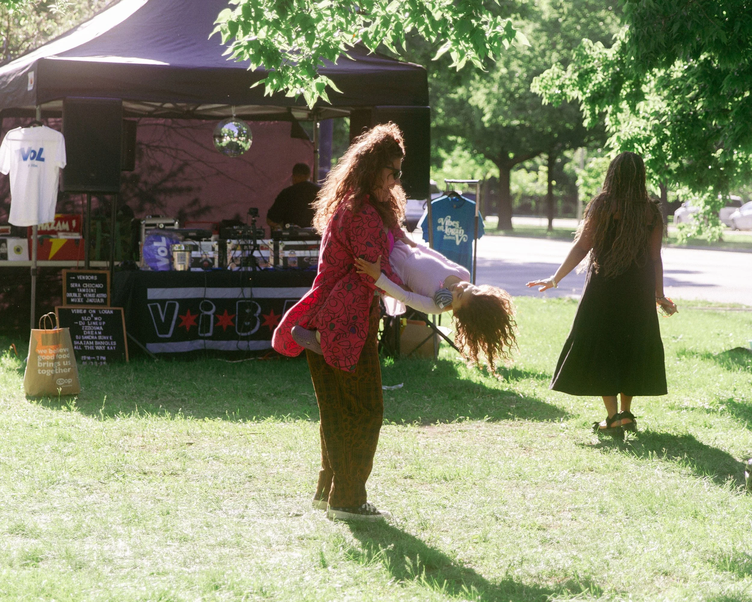 Women dancing outdoors in a park with a DJ booth and DJ equipment under a black canopy, with trees and a sidewalk in the background.