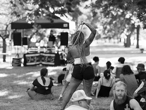 A woman dancing with a hula hoop in an outdoor park setting, surrounded by seated spectators and a stage in the background.