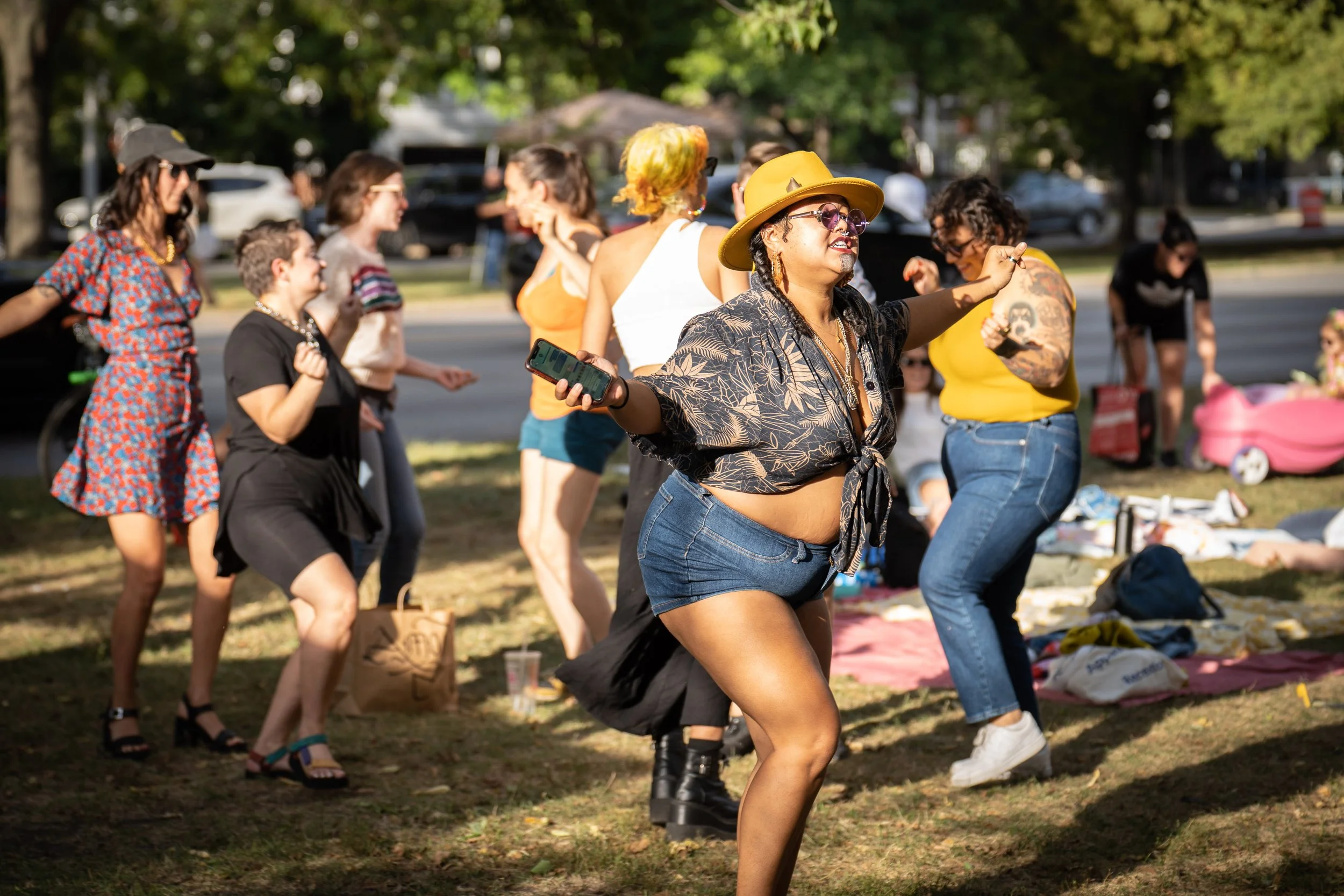Group of people dancing and socializing outdoors in a park, with trees and cars in the background.