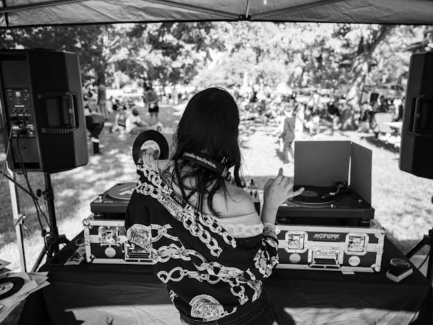 A woman DJ is performing at an outdoor event, with turntables and speakers set up under a canopy, in a park with trees and people in the background.