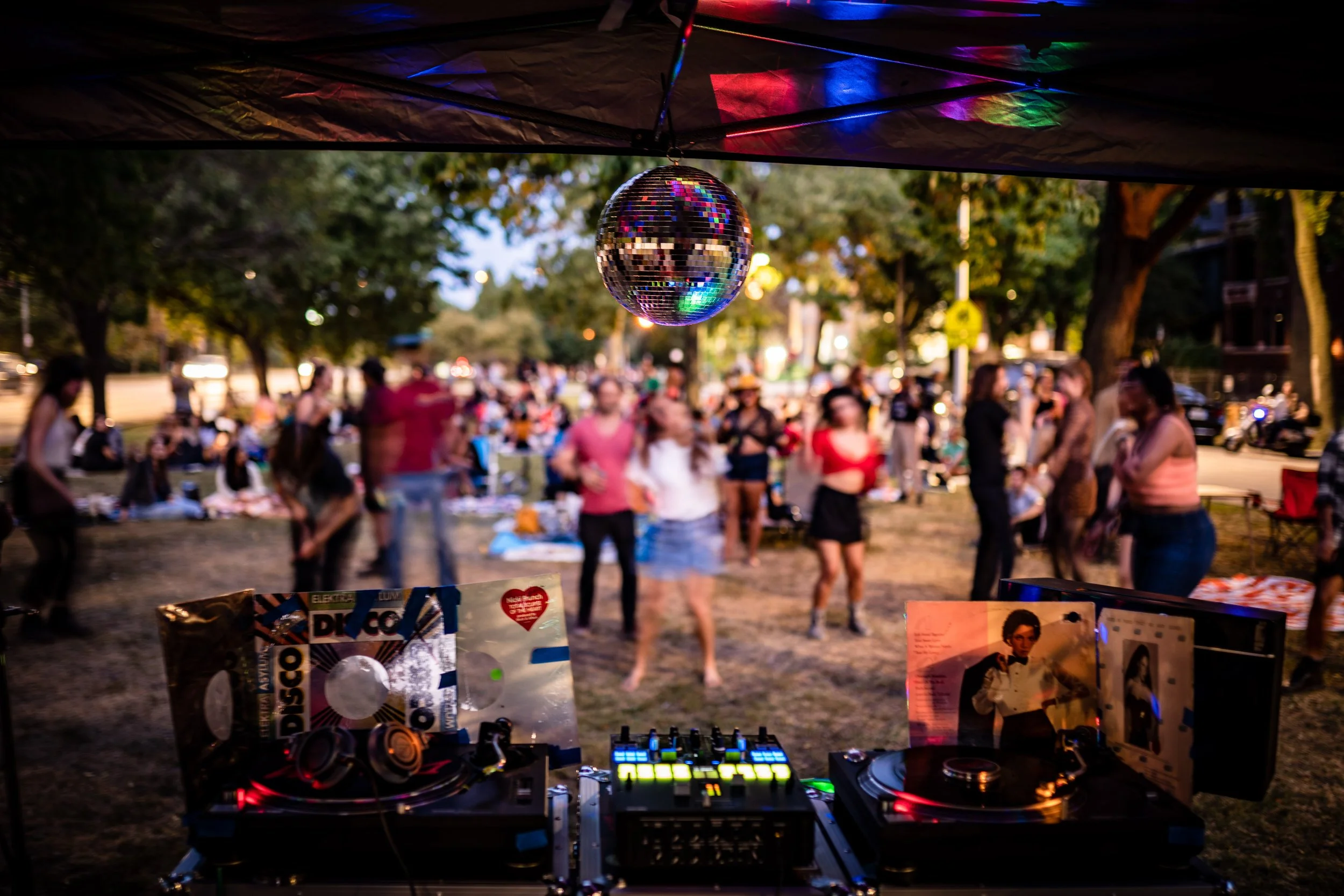 A DJ setup with turntables and electronic equipment in front of a crowd at an outdoor event during dusk, with a disco ball hanging from the ceiling, reflecting colorful lights. People are gathered in the background, some dancing and socializing under trees.