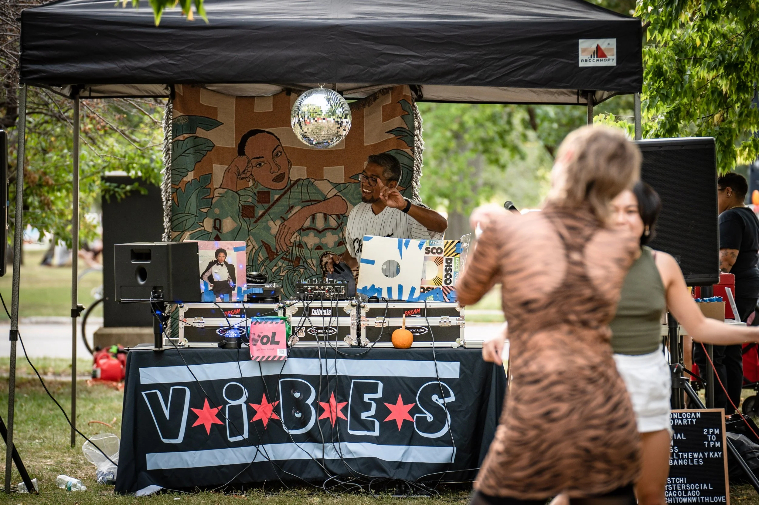 A DJ performing at an outdoor event with a black canopy tent, a colorful tapestry featuring a woman's face, and a disco ball hanging above the DJ. People are dancing in front of the setup in a park-like setting with green trees.