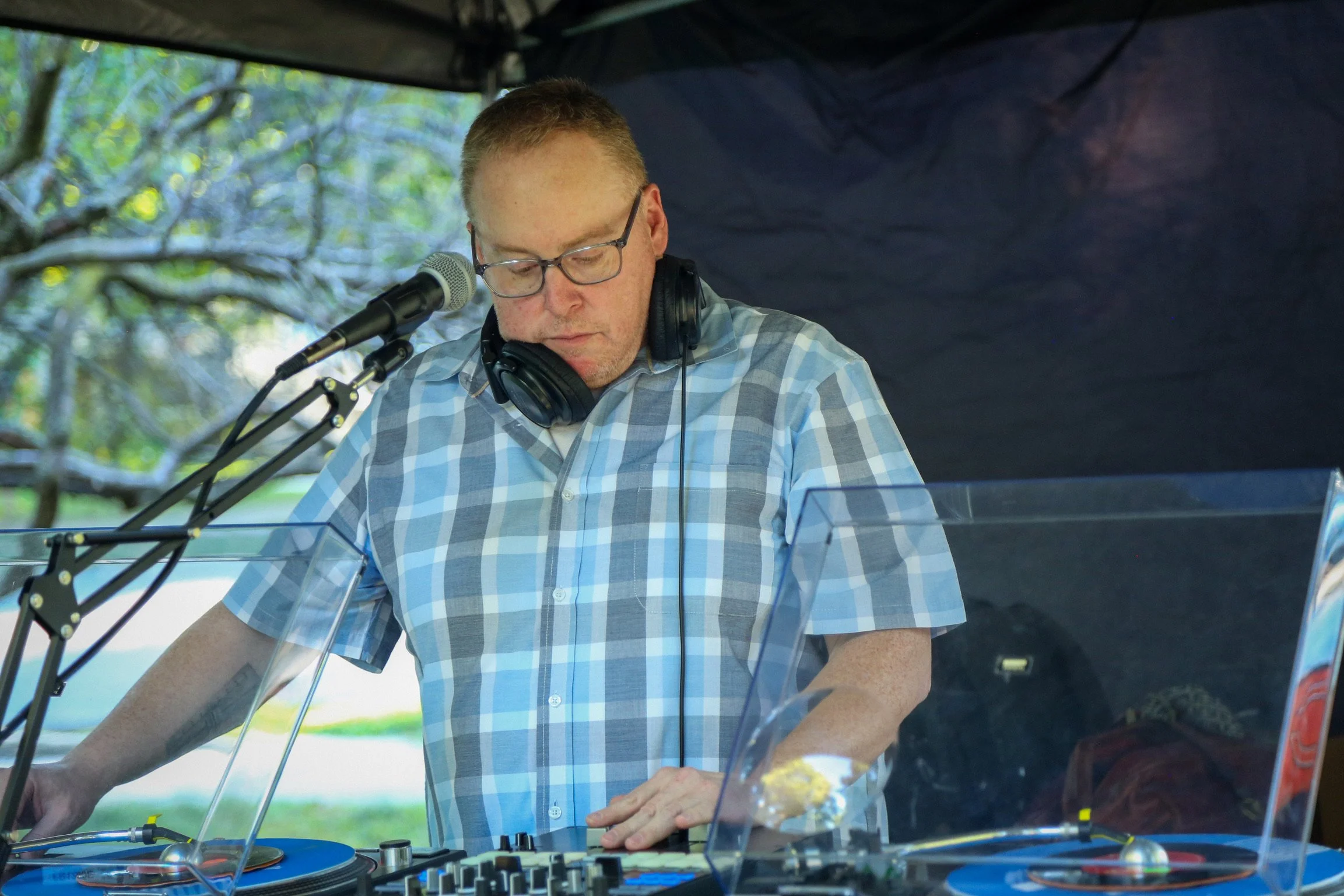 A man with glasses and headphones wearing a plaid shirt, operating DJ equipment outdoors under a canopy, with trees in the background.