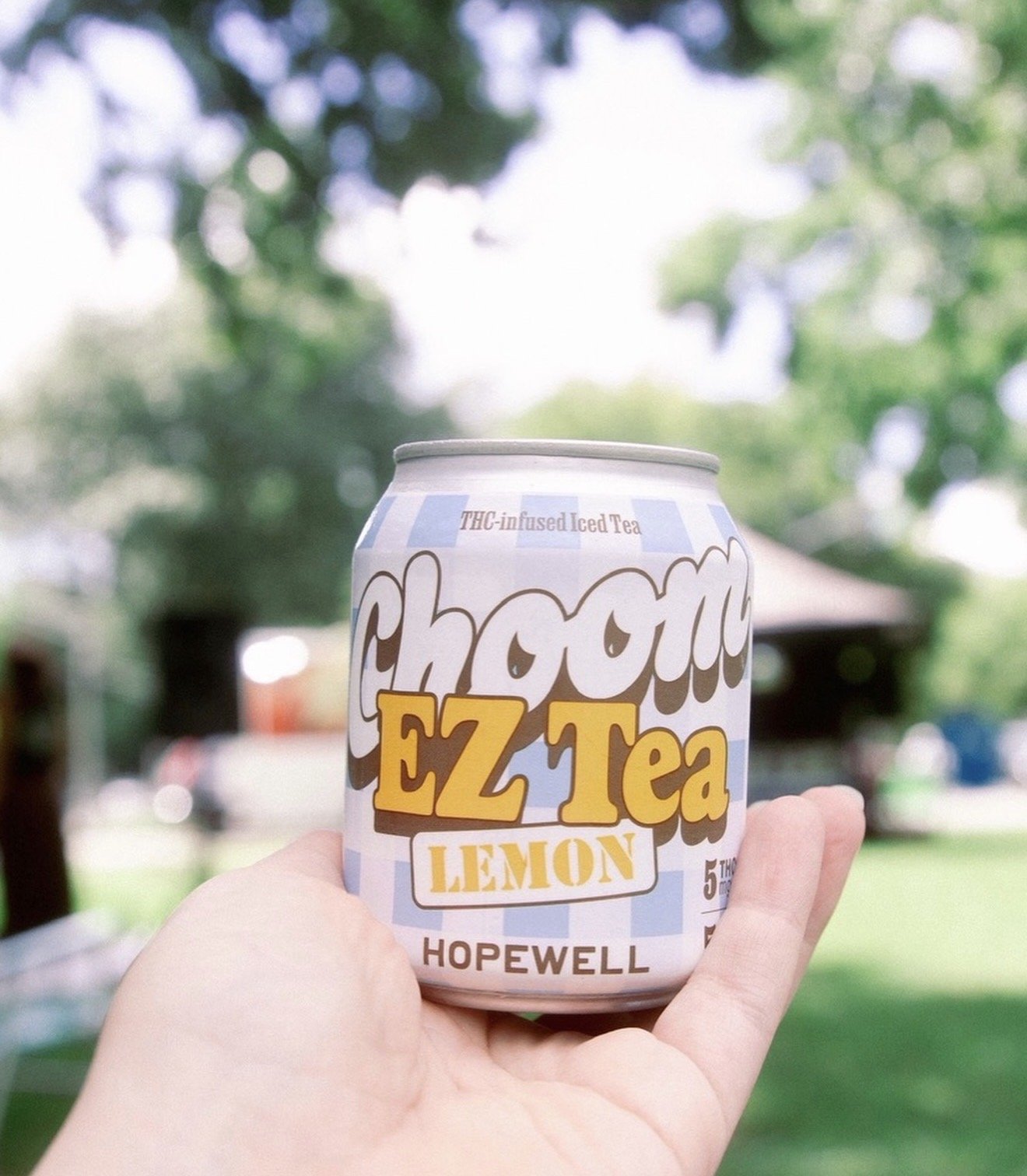A can of Choom EZ Tea Lemon held outdoors with a blurred background of green trees and structures.