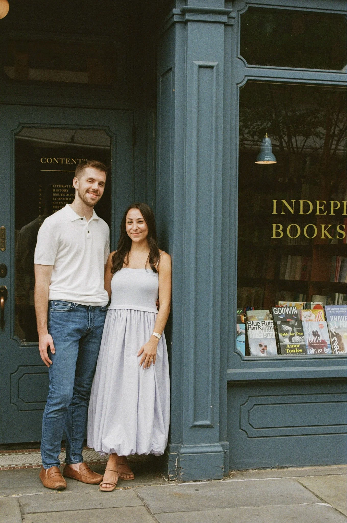A young man and woman stand outside a bookstore, smiling at the camera. The bookstore has a blue exterior and visible display of books in the window.