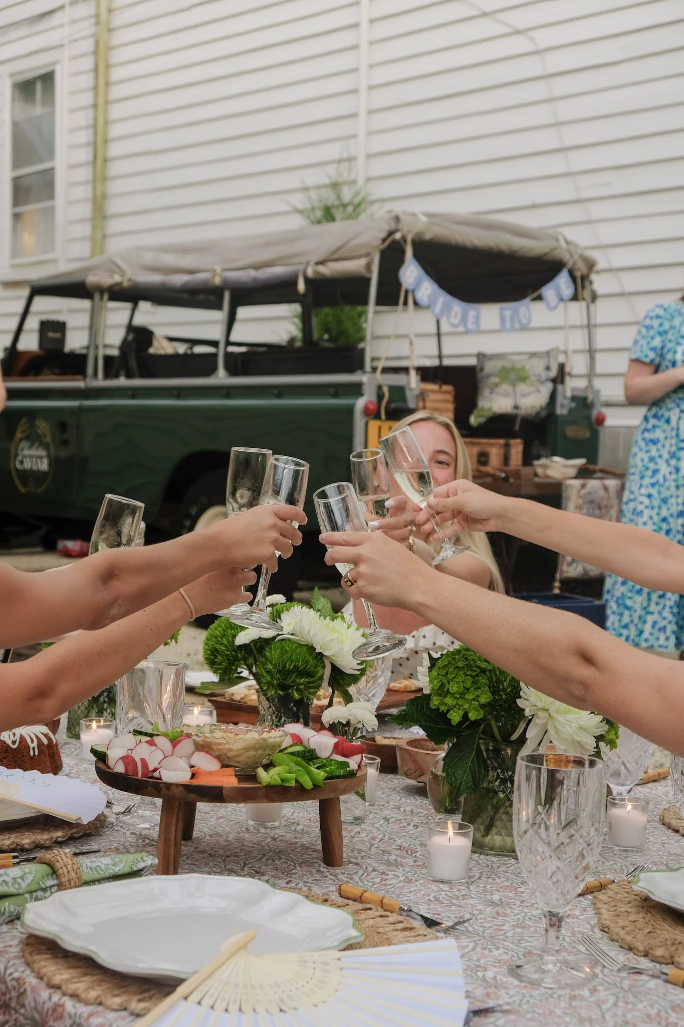 People celebrating outdoors with clinking champagne glasses at a dinner table decorated with flowers, candles, and various foods.
