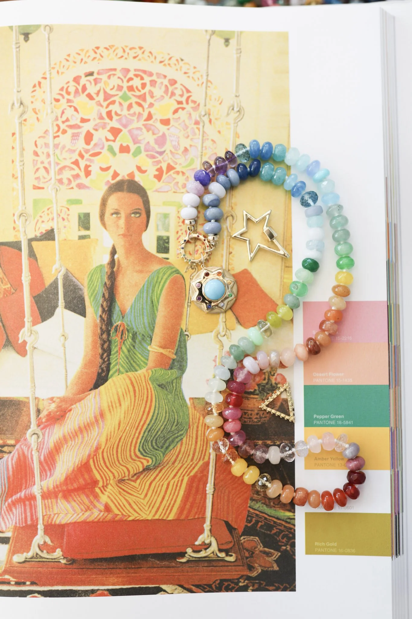 A jewelry display with colorful bead necklaces and a silver star and triangle pendant on top of a magazine page featuring a woman in a colorful striped dress sitting in a swing.